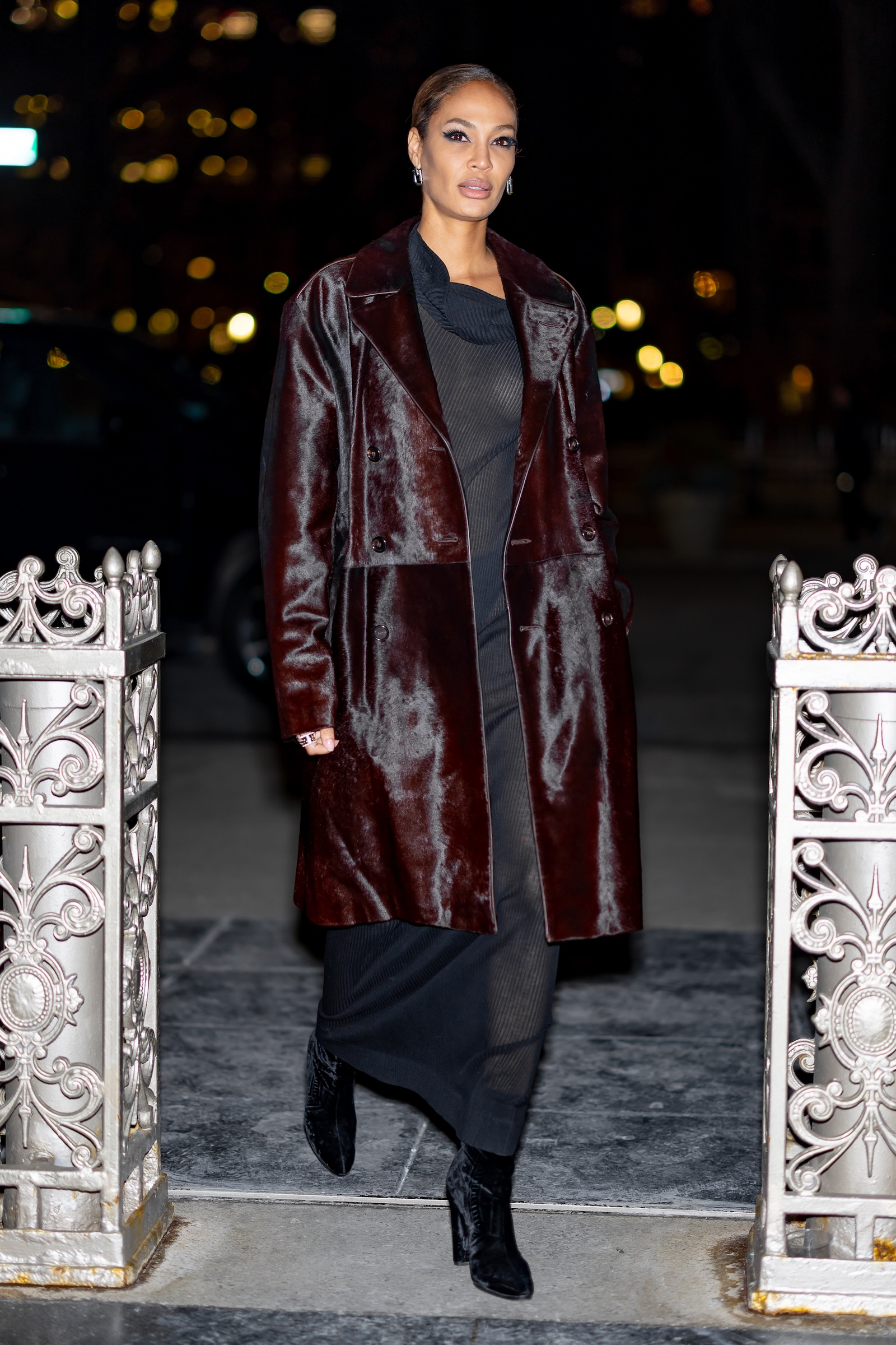 Joan Smalls w czarnej sukience maxi, welurowych czarnych botkach i brązowym płaszczu.