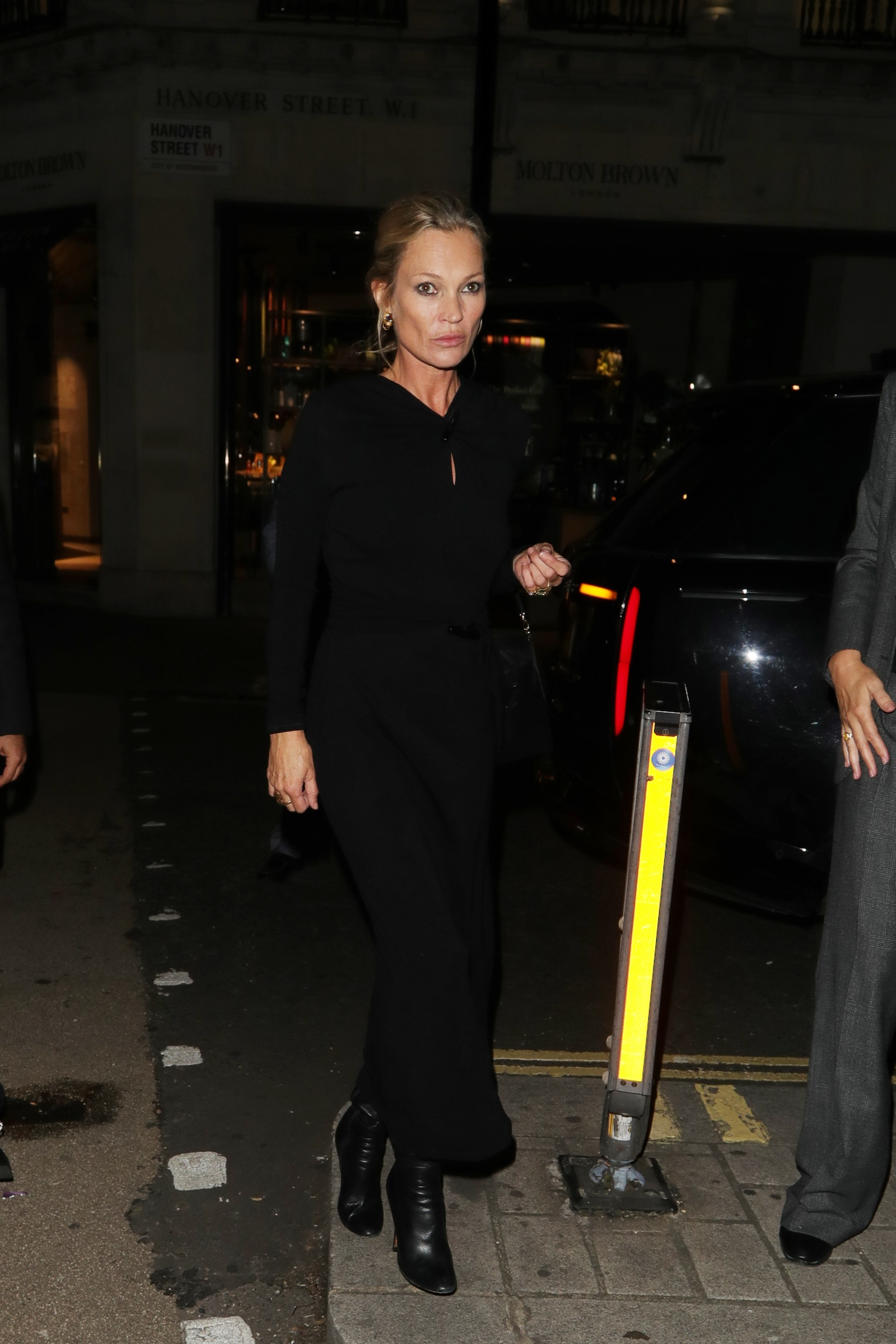 Kate Moss w długiej czarnej sukience i skórzanych czarnych kozaki na ulicy, wieczorna sceneria.