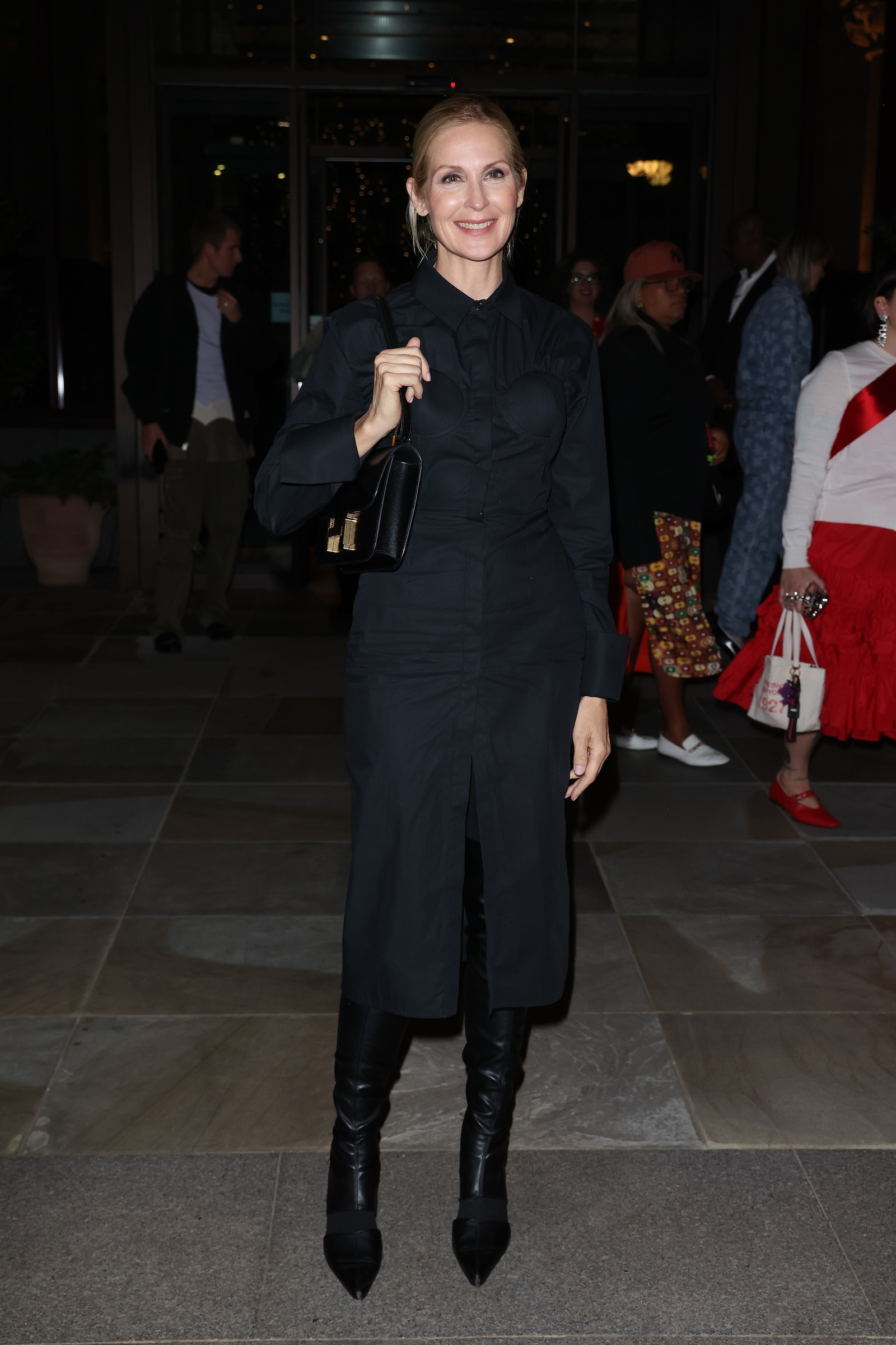 Kelly Rutherford w czarnej sukience koszulowej, czarnych kozakach za kolano i czarnej torebce na ramię.
