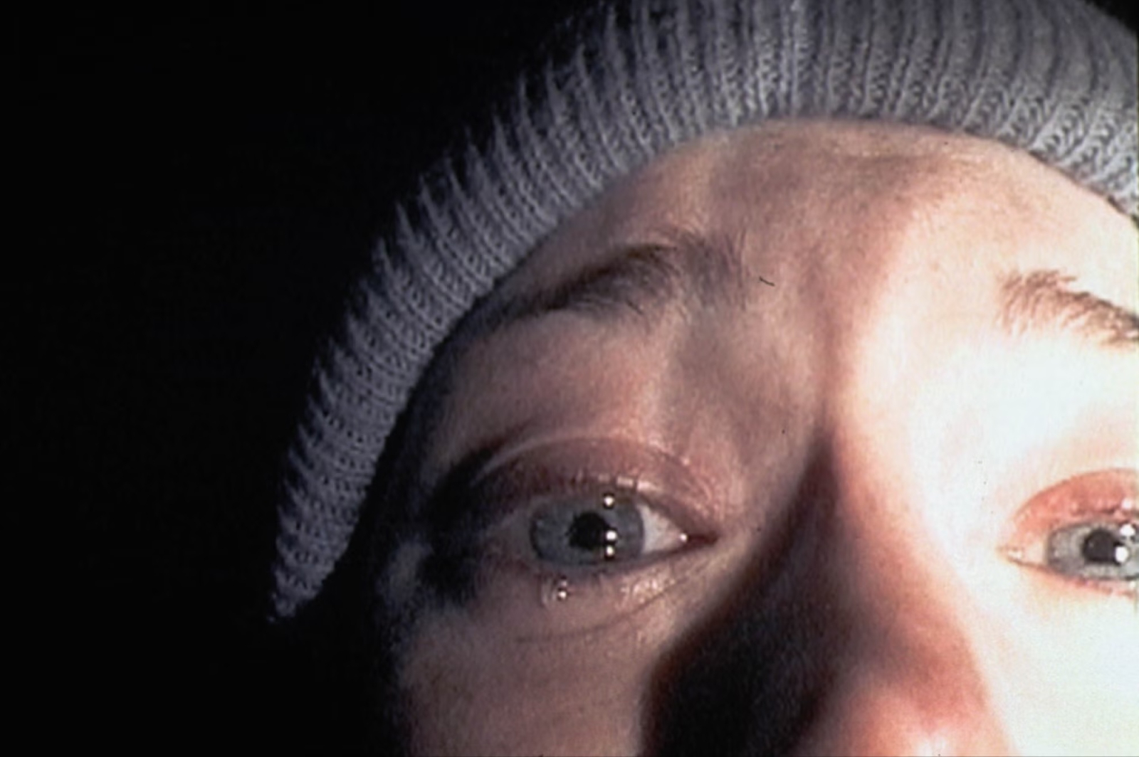 Blisko ujęcie twarzy postaci w kadrze z horroru „Blair Witch Project”, z wyraźnym przerażeniem w oczach.