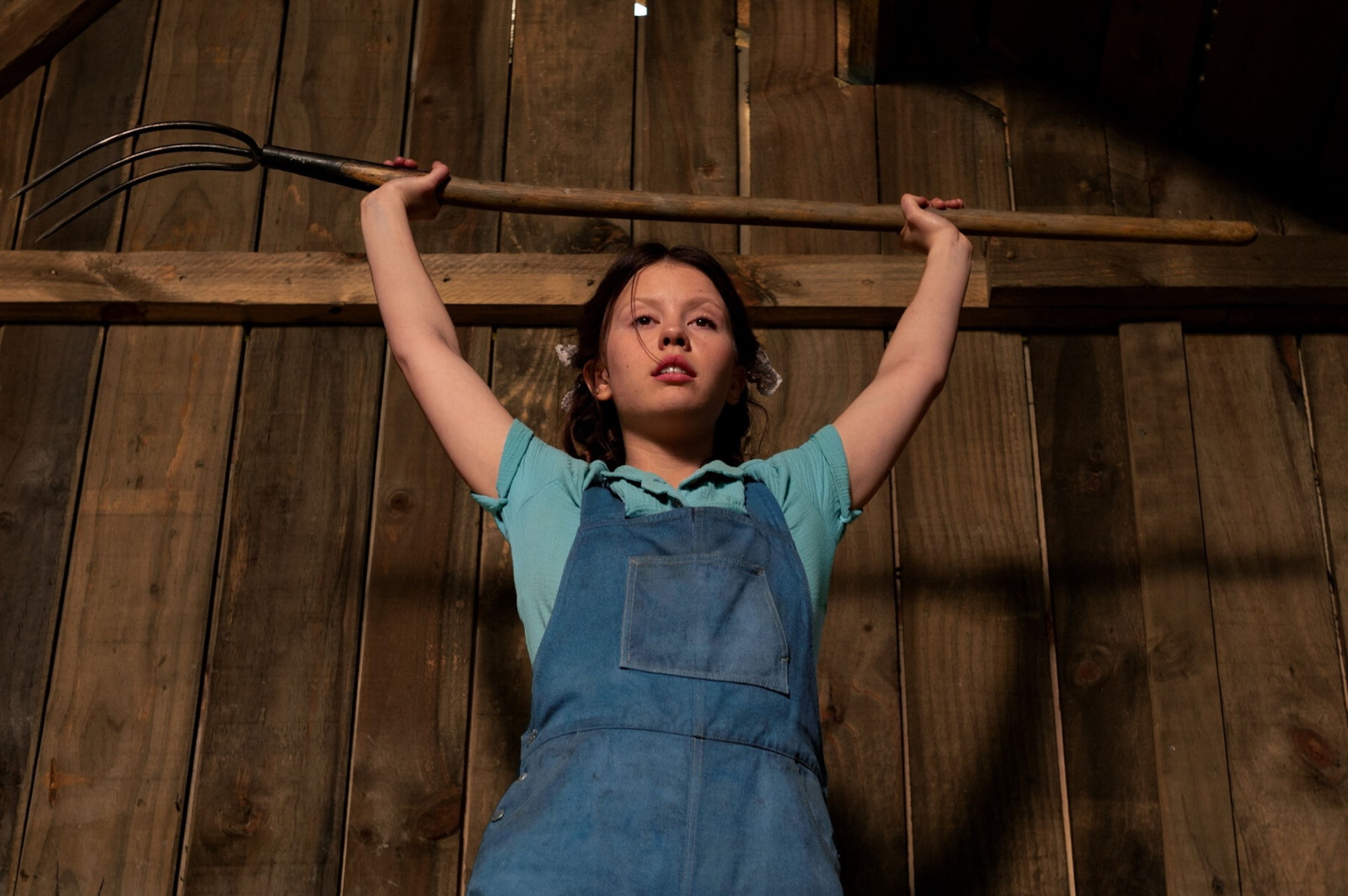 Mia Goth z filmu grozy „Pearl” trzyma widły, stojąc w drewnianym wnętrzu.