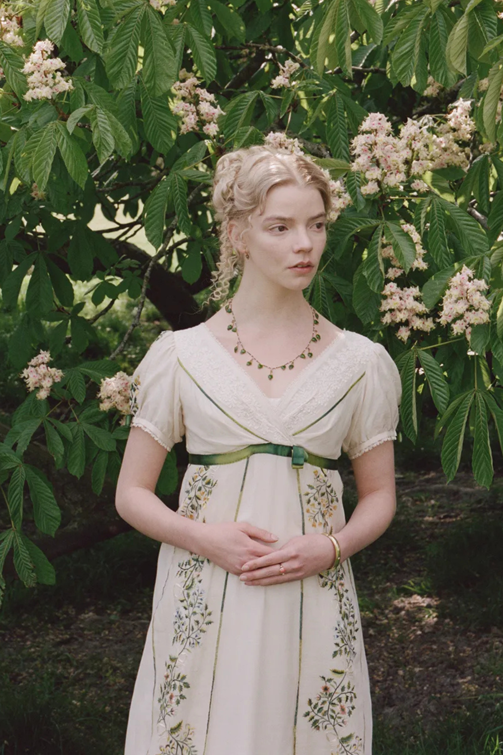 Anya Taylor-Joy w filmie „Emma” w jasnej sukni, stojąca wśród kwitnących gałęzi drzewa.