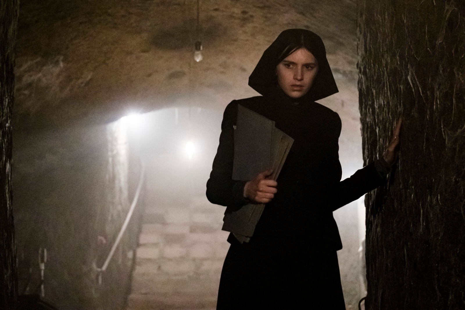 Nell Tiger Free w filmie „Omen: Początek” stoi w ciemnym, kamiennym korytarzu, trzymając teczki.