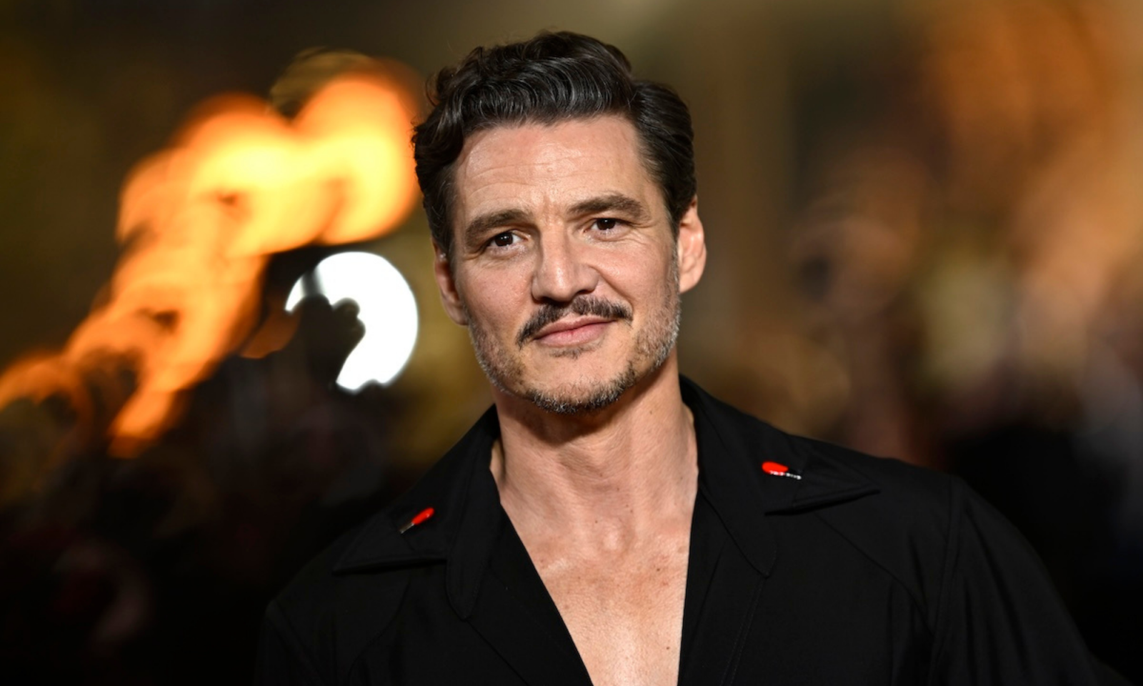 Pedro Pascal z uśmiechem, ubrany w czarną koszulę, na tle rozmytych świateł.