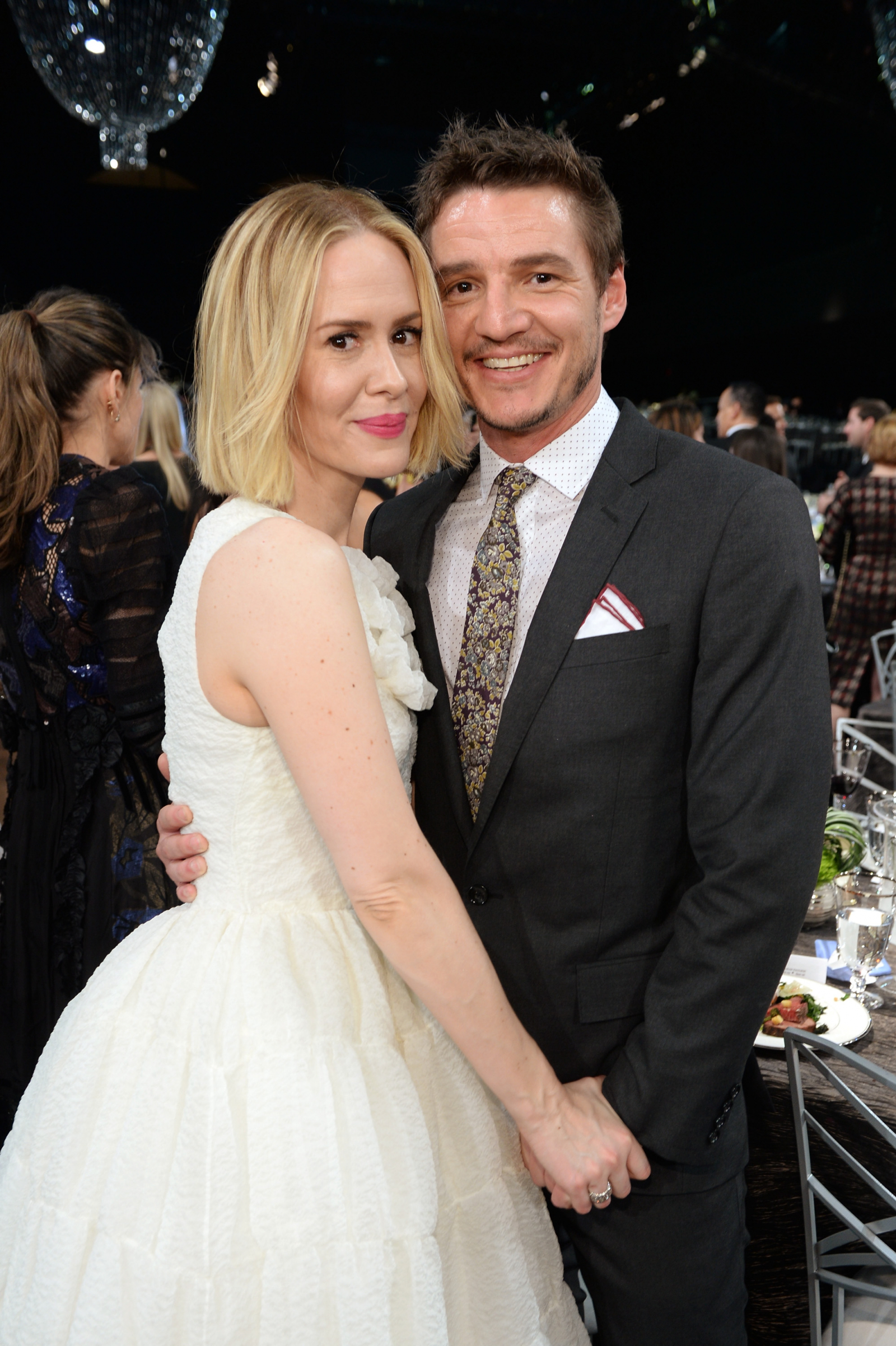 Sarah Paulson i Pedro Pascal pozują razem w eleganckich strojach, w tle sala z gośćmi i dekoracjami.