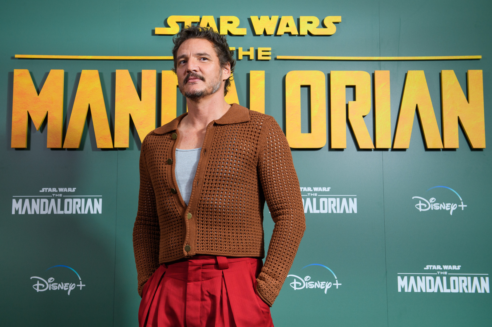 Pedro Pascal na premierze "The Mandalorian" z logo Star Wars w tle, ubrany w sweter i czerwone spodnie.