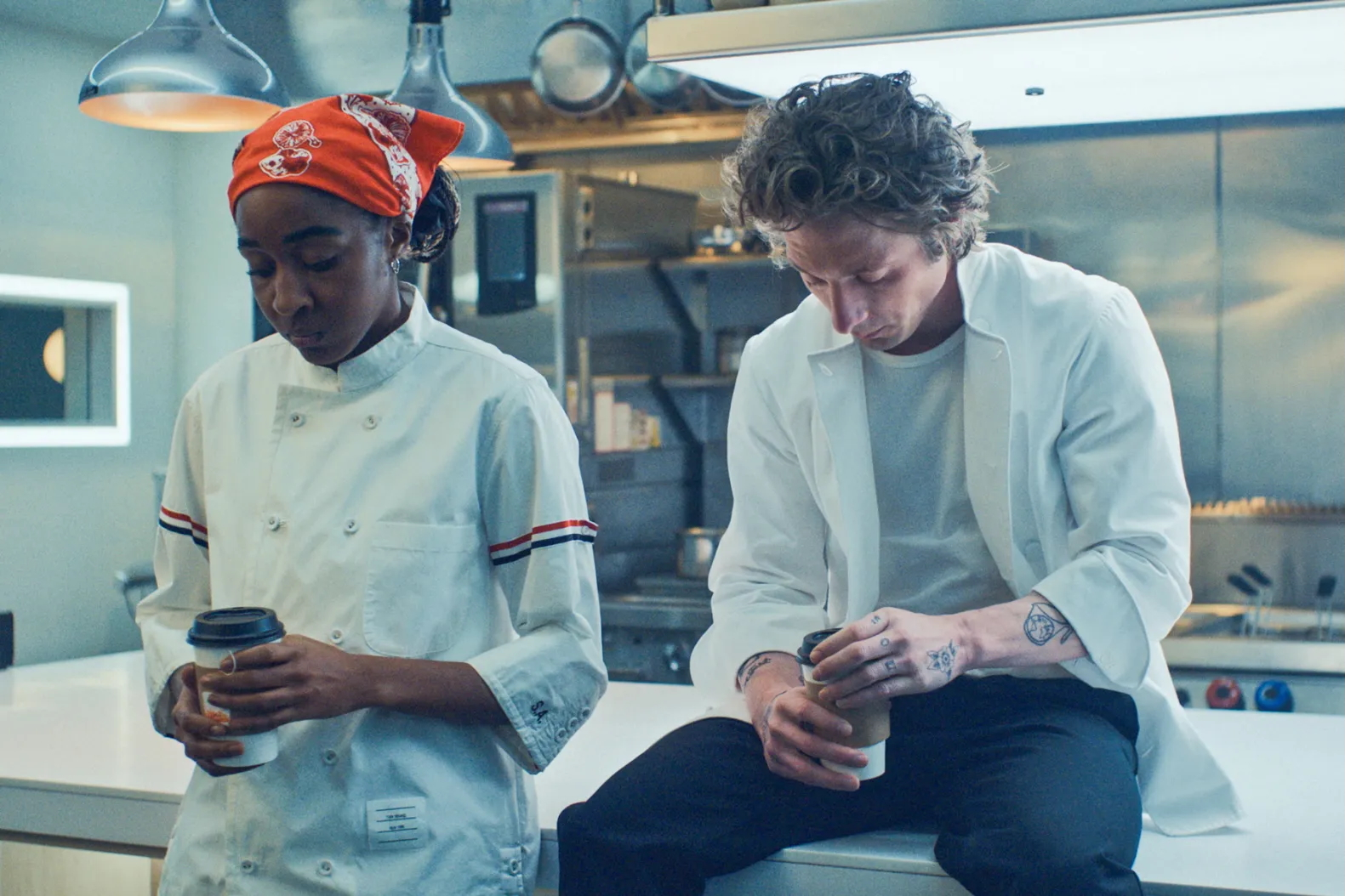Ayo Edebiri i Jeremy Allen White w kuchni, siedzą przy blacie, trzymając kubki z kawą. Atmosfera napięta.