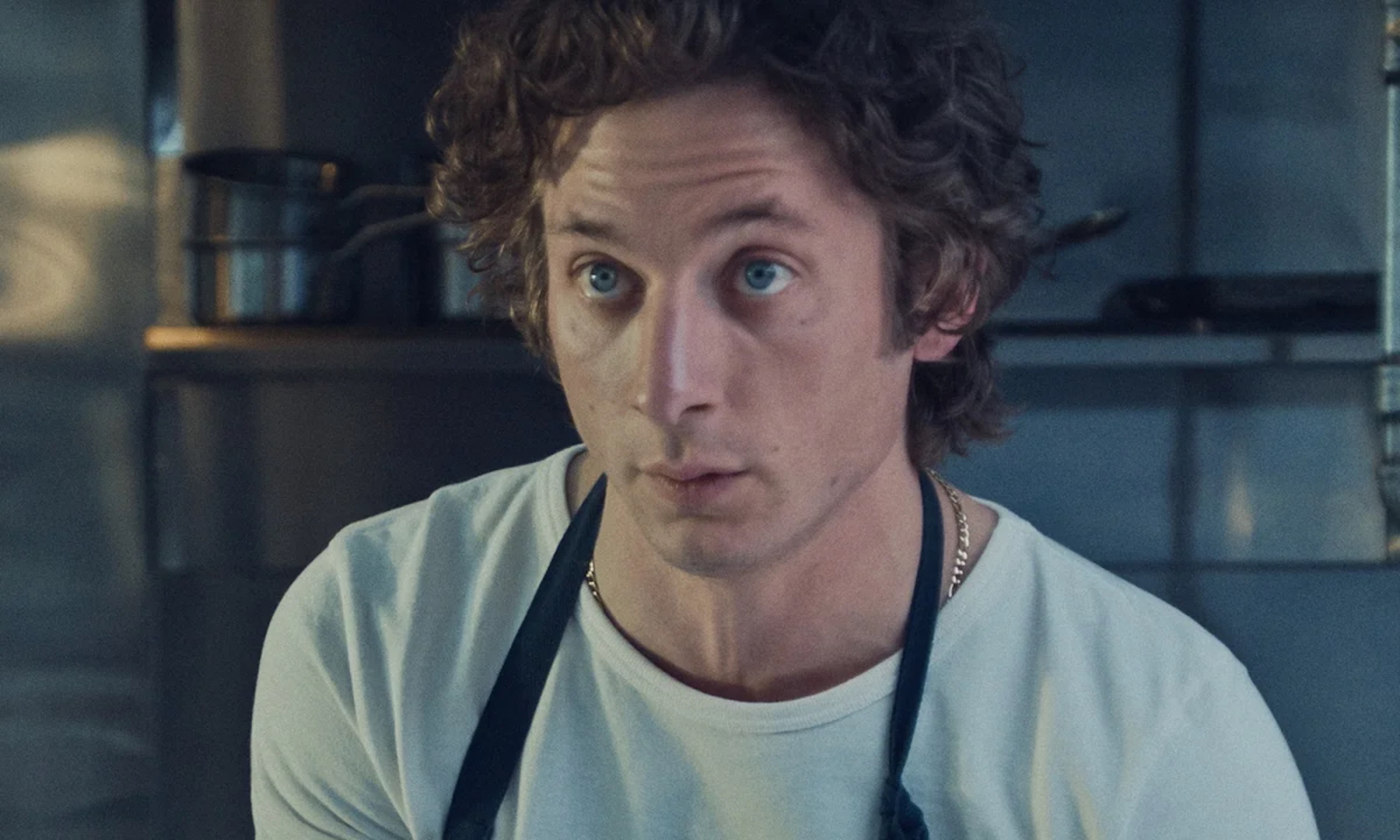 Jeremy Allen White w kuchni, w białym t-shircie i fartuchu, z intensywnym spojrzeniem, przygląda się czemuś z zainteresowaniem.
