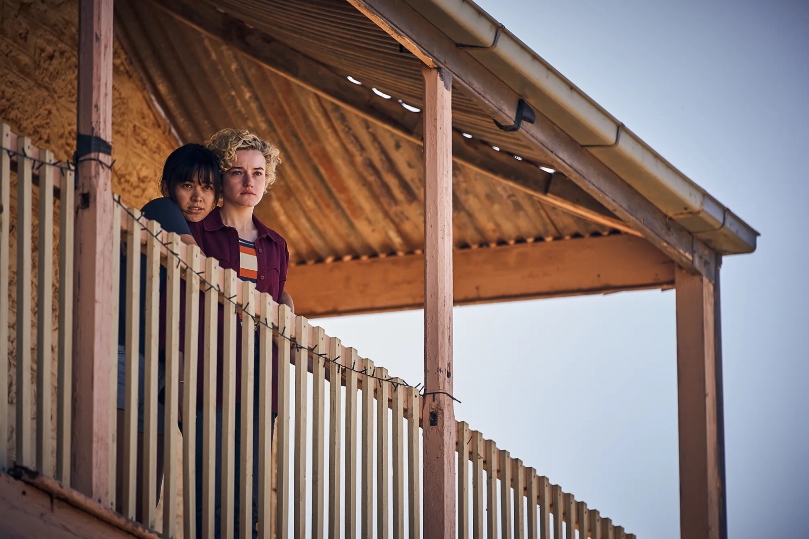 Jessica Henwick i Julia Garner w filmie „The Royal Hotel” stoją na balkonie, spoglądając w dal.