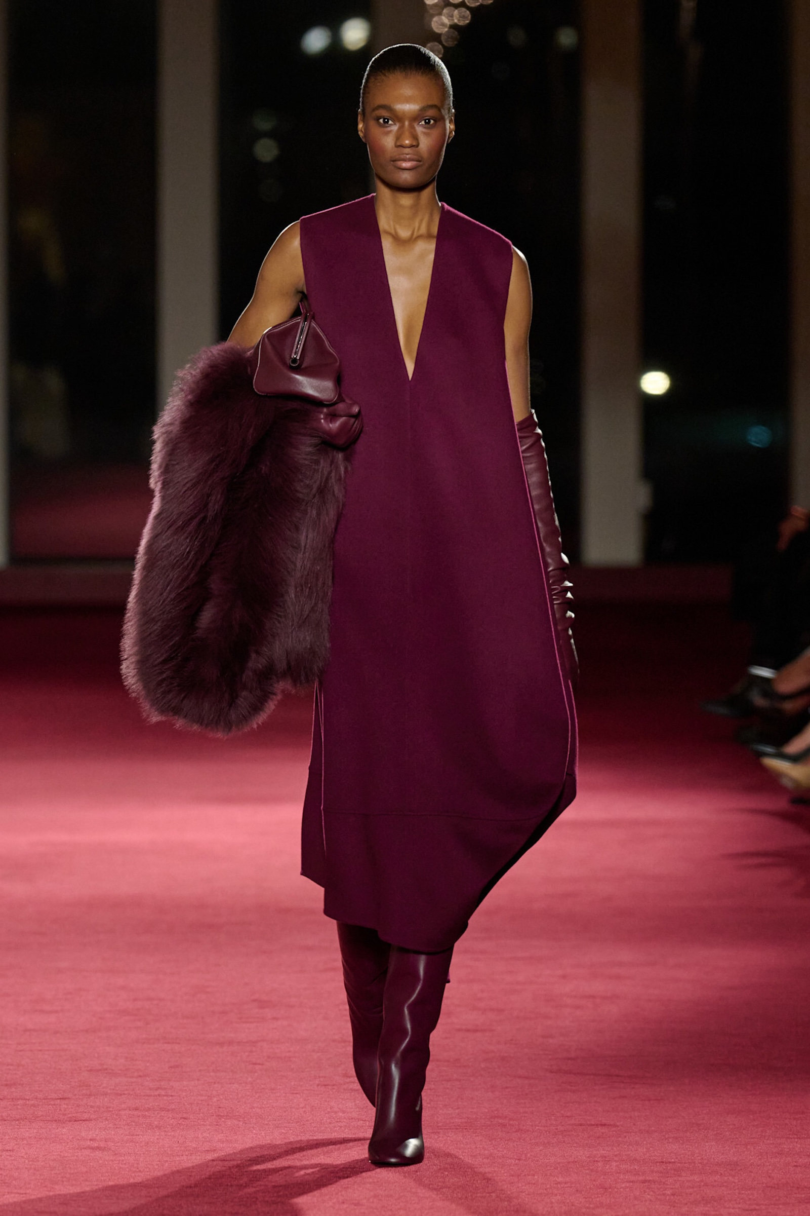 Modelka w burgundowych kozakach i luźnej sukience, trzyma futrzany płaszcz, prezentując elegancki styl na czerwonym tle.