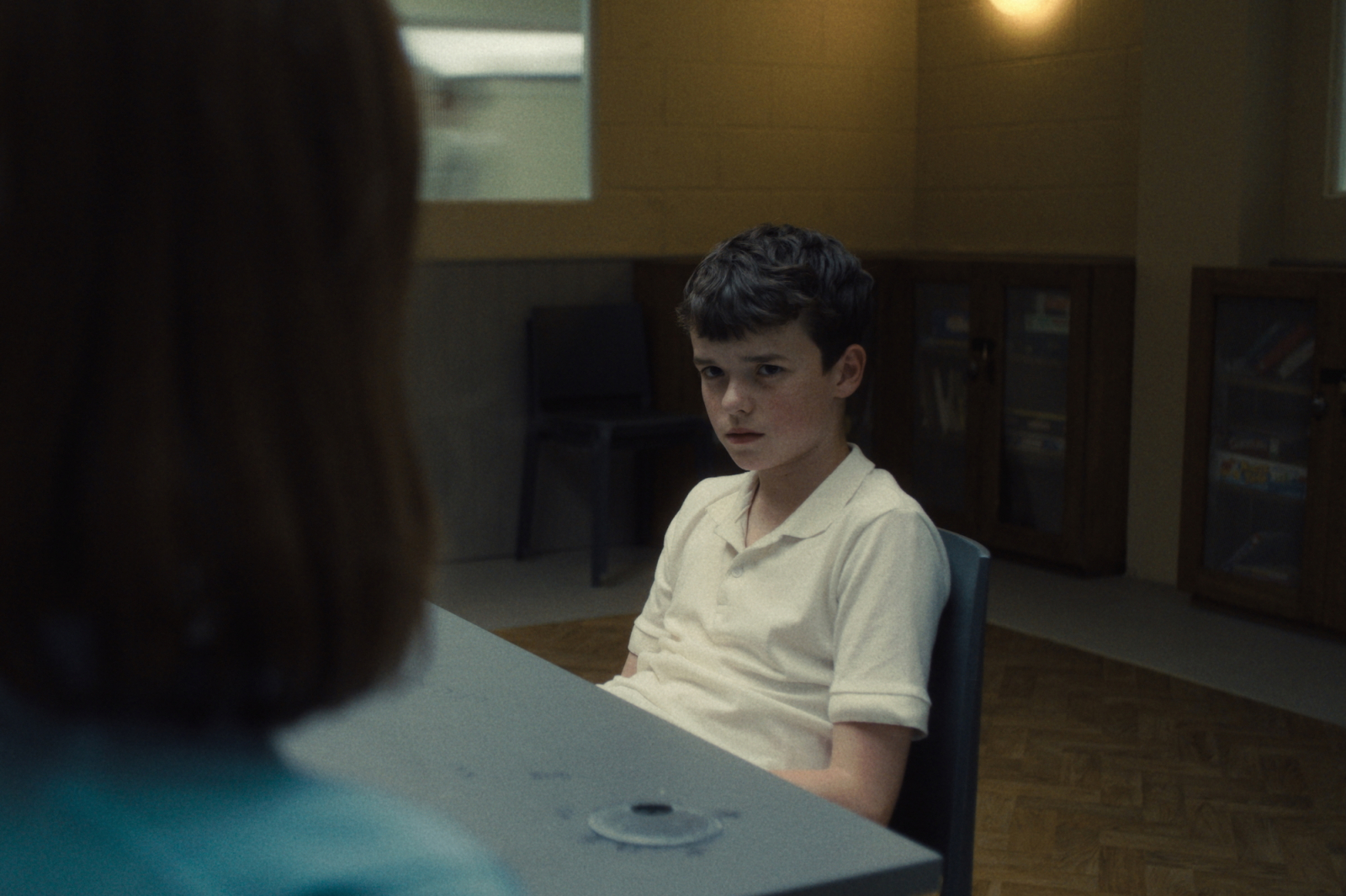 Owen Cooper w trakcie rozmowy w serialu Netflix „Dojrzewanie”, siedzi przy biurku z poważnym wyrazem twarzy.