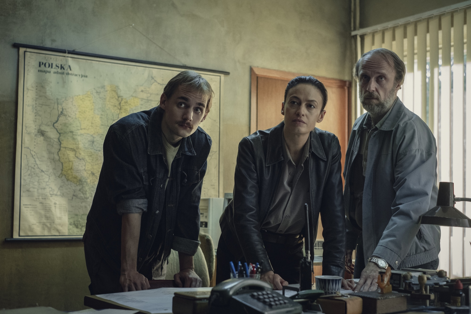 Magdalena Różczka w scenie z serialu Netflix „Rojst”, stojąc z dwoma mężczyznami w biurze z mapą Polski w tle.