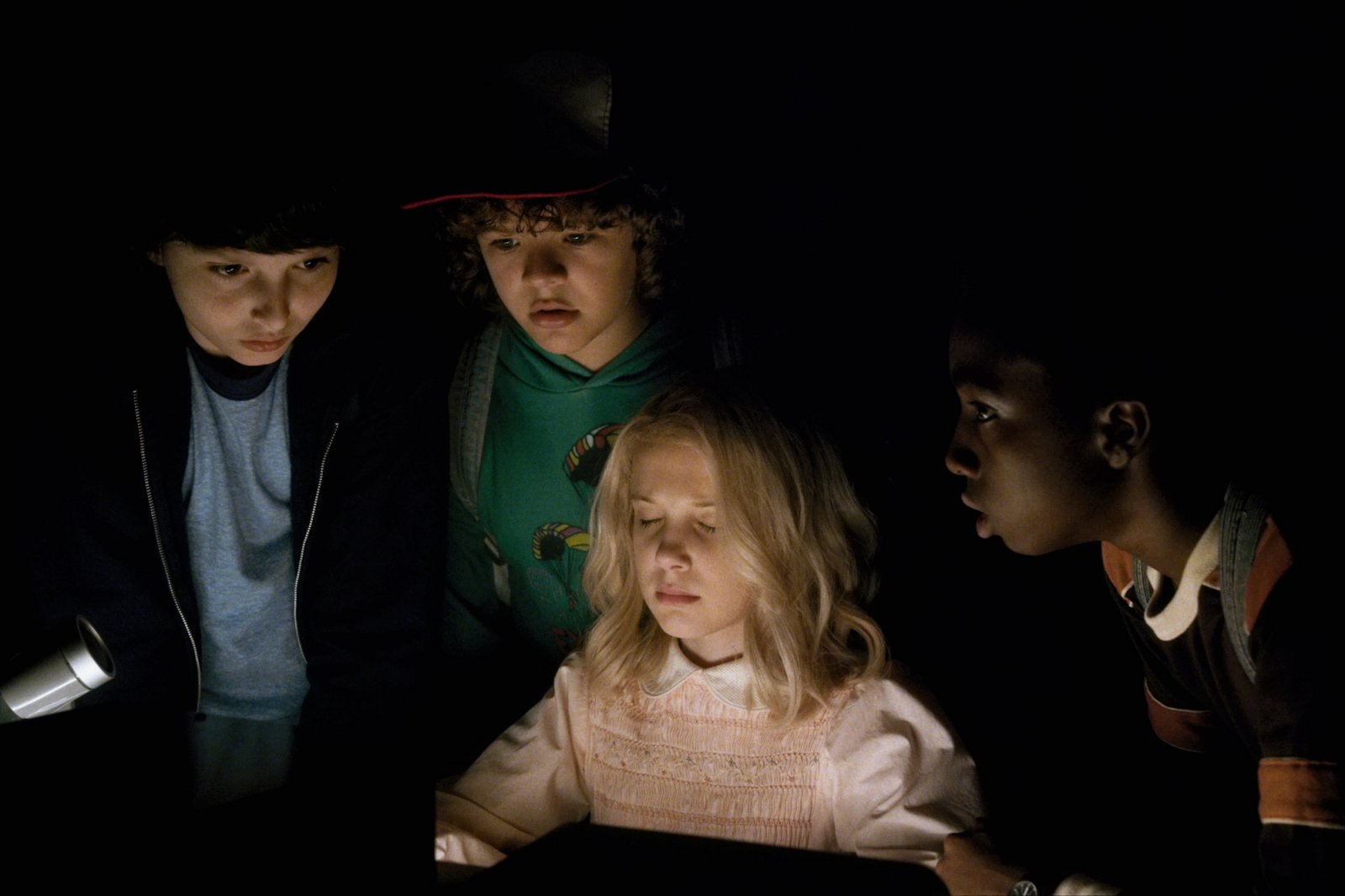 Czwórka dzieci z serialu Netflix „Stranger Things” w ciemności, skupiona na świecącym obiekcie.