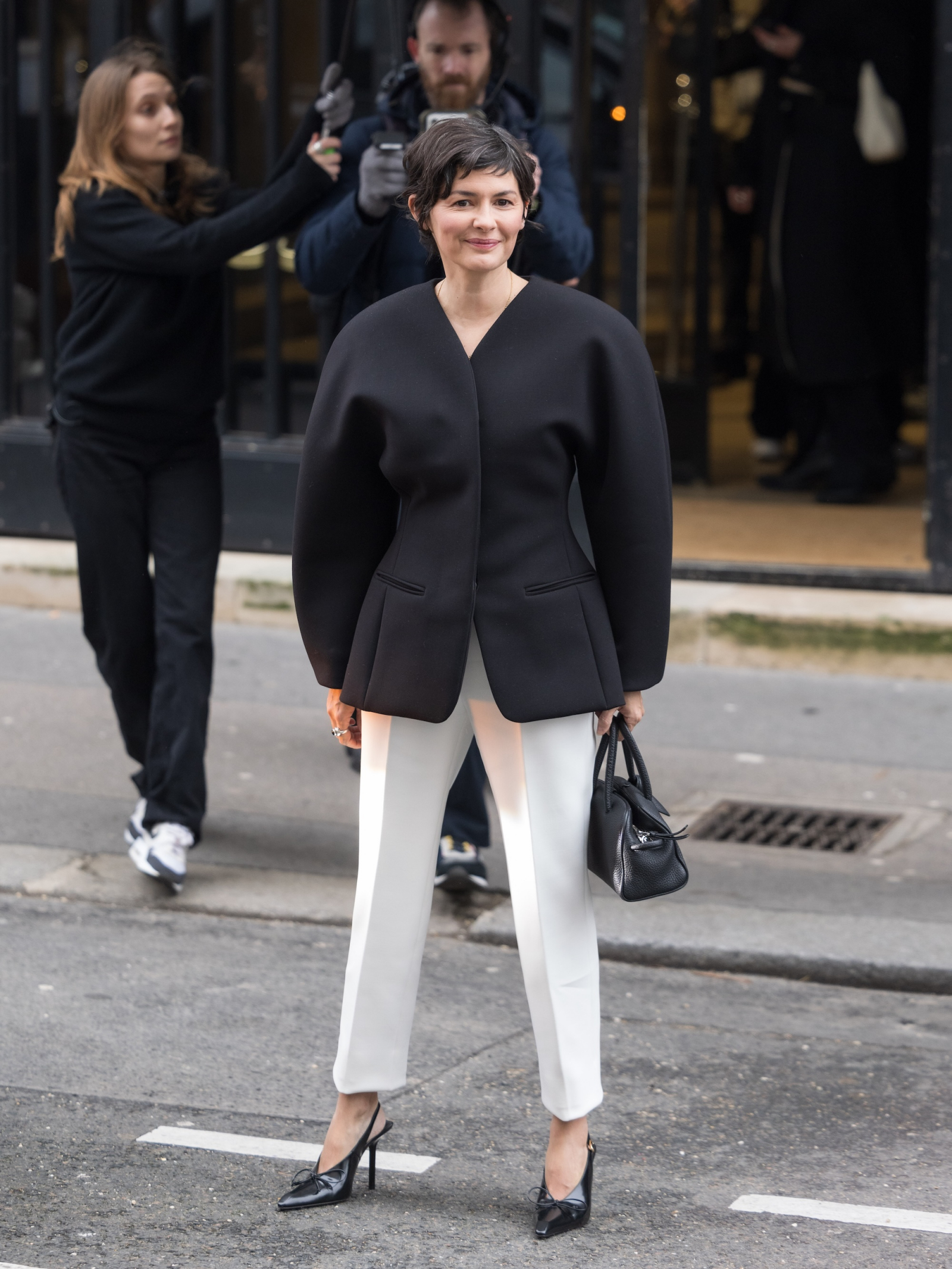 Audrey Tautou w eleganckim czarnym płaszczu i białych spodniach damskich, pozująca na ulicy.