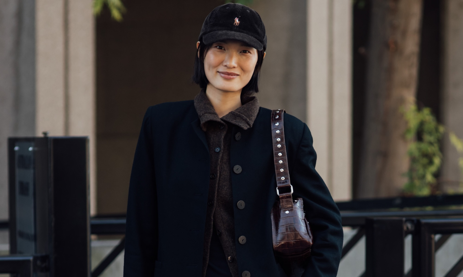 Kobieta w eleganckim czarnym płaszczu z brązowym sweterkiem i czapką, trzyma małą torebkę. Tło to urbanistyczne otoczenie.