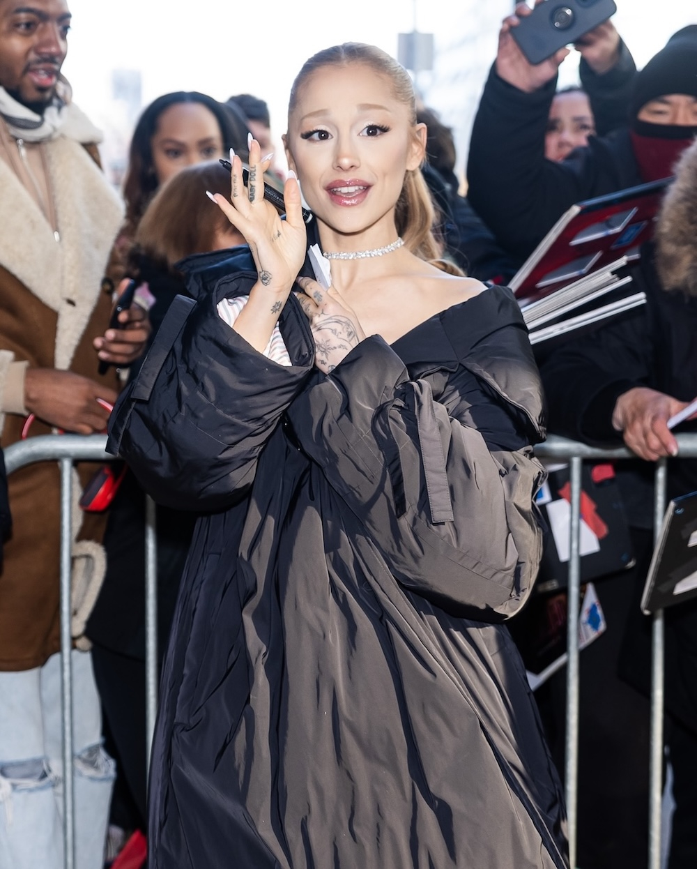 Ariana Grande w długiej puchowej czarnej kurtce damskiej, pozdrawia fanów na ulicy.