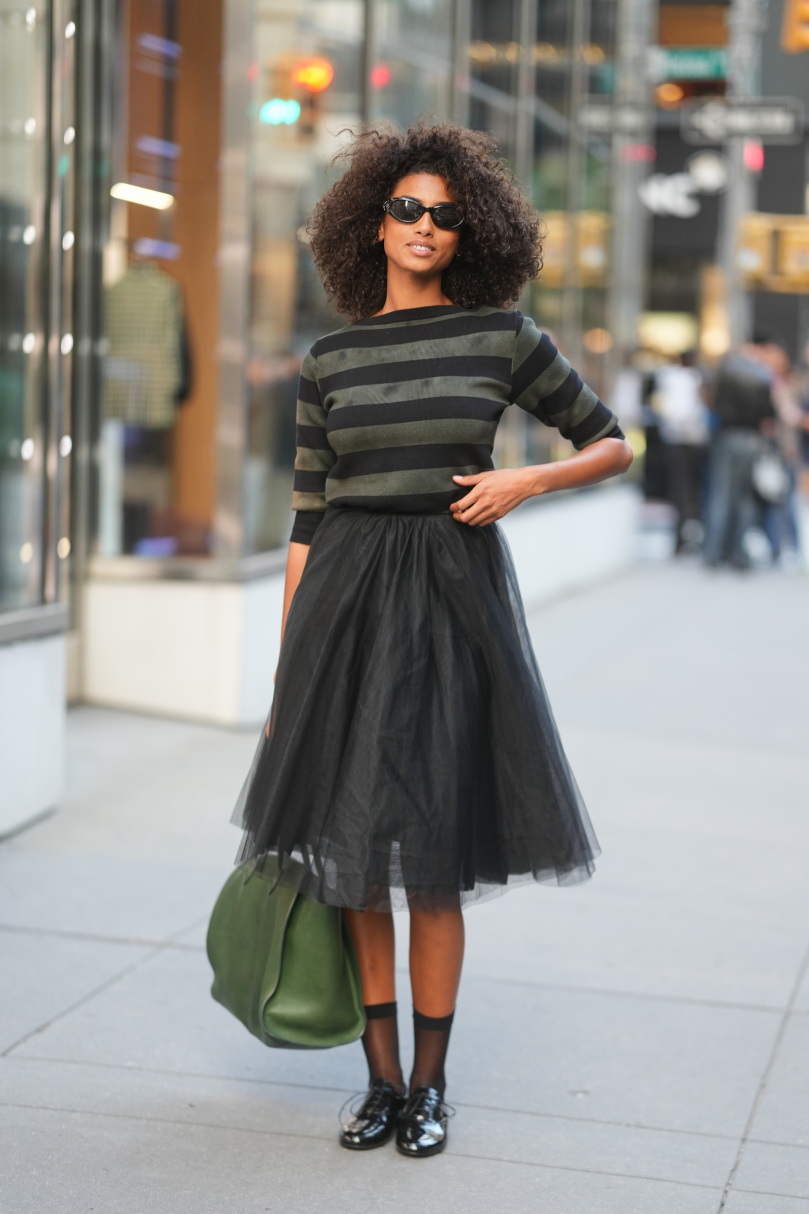 Imaan Hammam w czarno-zielonym stroju, skórzane czarne oxfordki damskie i zielona torba.