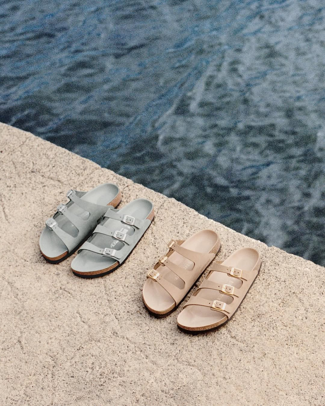 Skórzane klapki damskie Birkenstock Florida w kolorze szarym i beżowym, leżące na kamieniu przy wodzie.