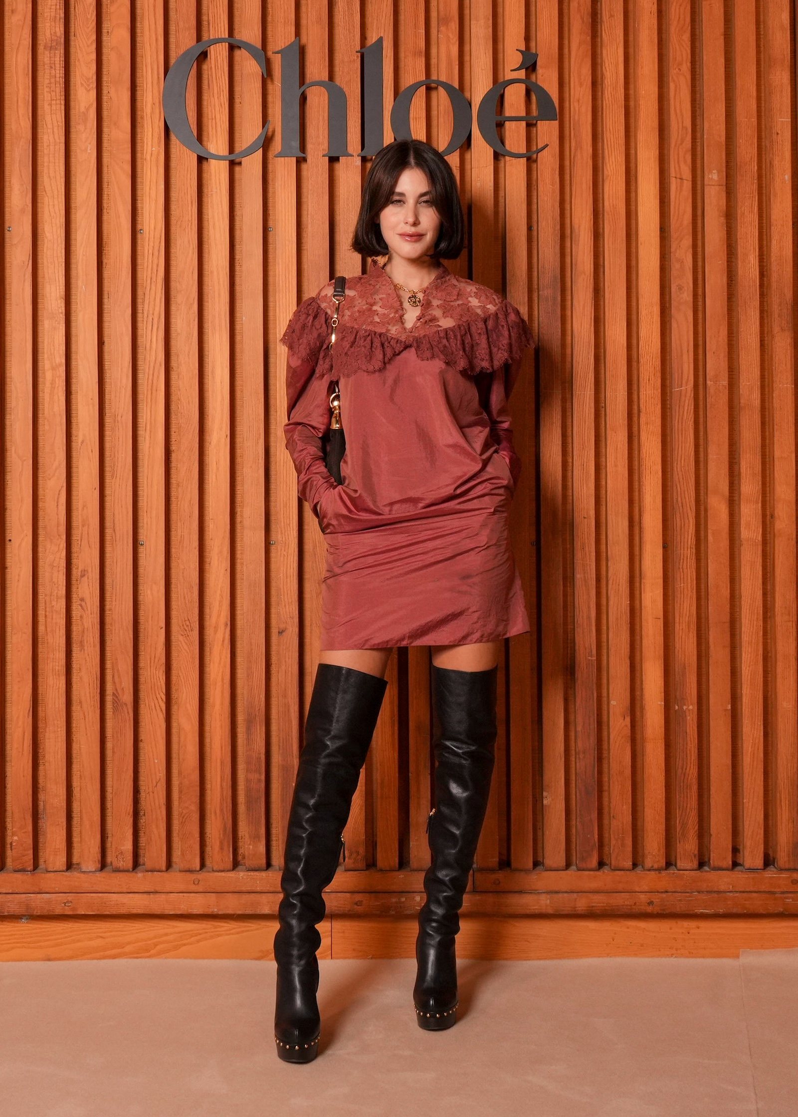 Kobieta w eleganckiej sukience mini w stylu boho, nosząca dopasowane skórzane kozaki na słupku, stoi przed drewnianą ścianą.