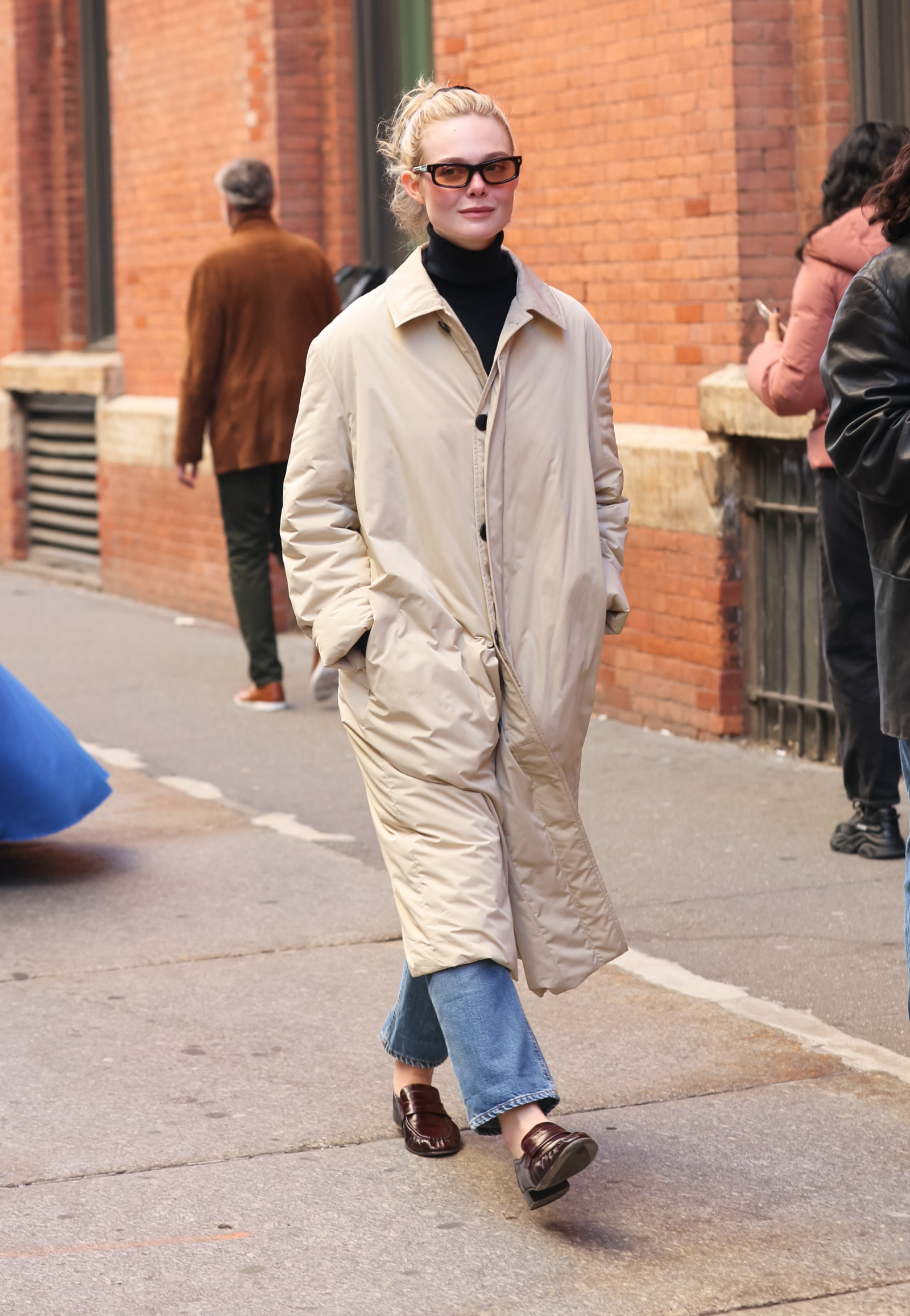 Elle Fanning w beżowym płaszczu i brązowych mokasynach damskich spaceruje po ulicy.