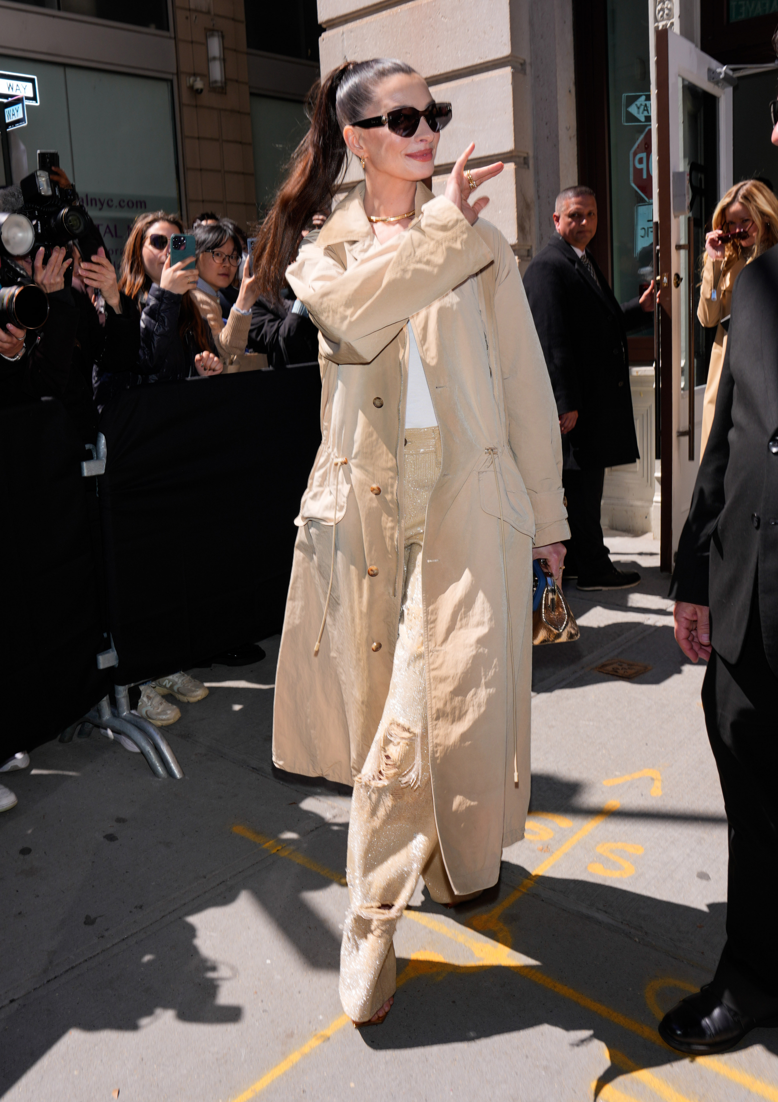 Anne Hathaway w beżowym płaszczu i jeansach z cekinami, wita fanów podczas wydarzenia.
