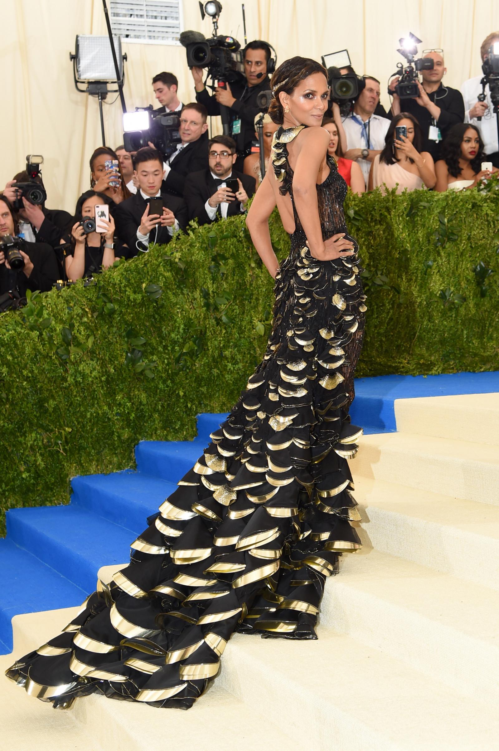 Halle Berry na MET Gala 2017 w efektownej, czarno-złotej sukni z falbanami, pozująca na schodach.