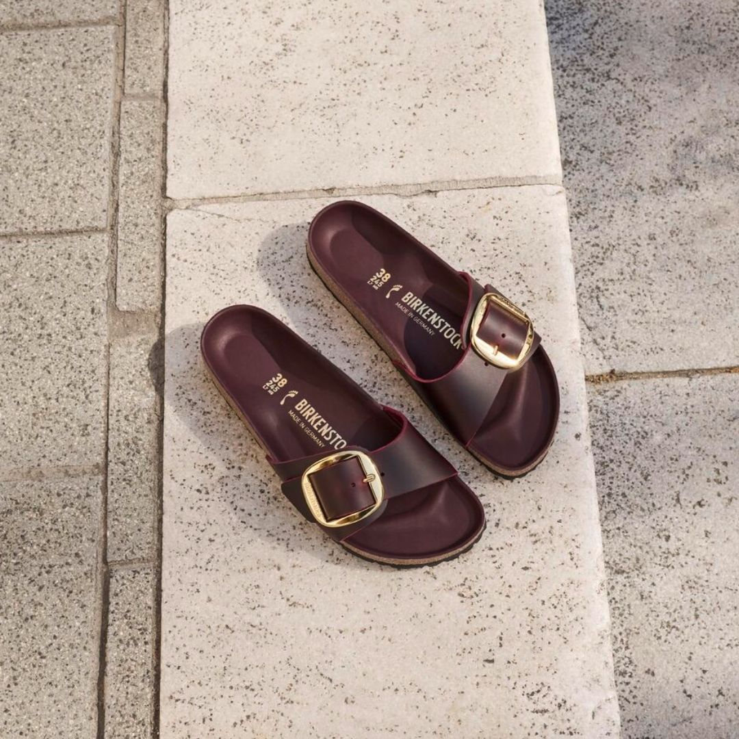Skórzane klapki damskie Birkenstock Madrid w ciemnym kolorze z dużą złotą klamrą, leżące na jasnym podłożu.