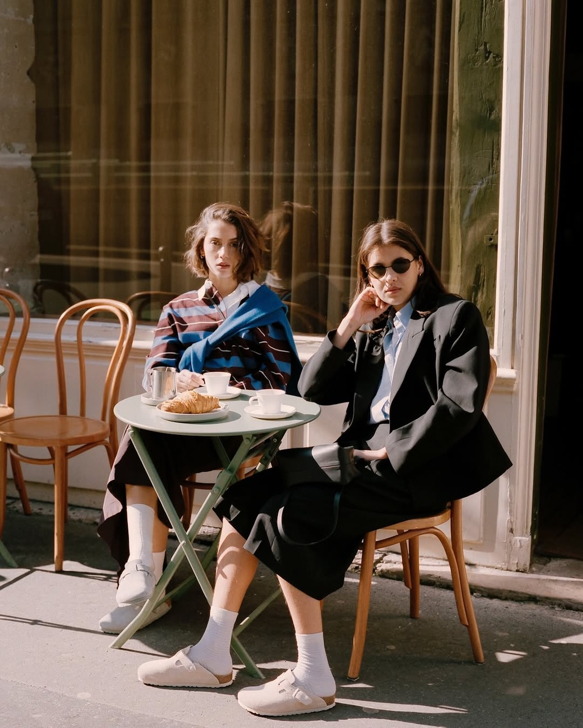 Dwie kobiety siedzą przy zielonym stole, noszą damskie klapki Birkenstock Boston, piją kawę i jedzą croissanty.