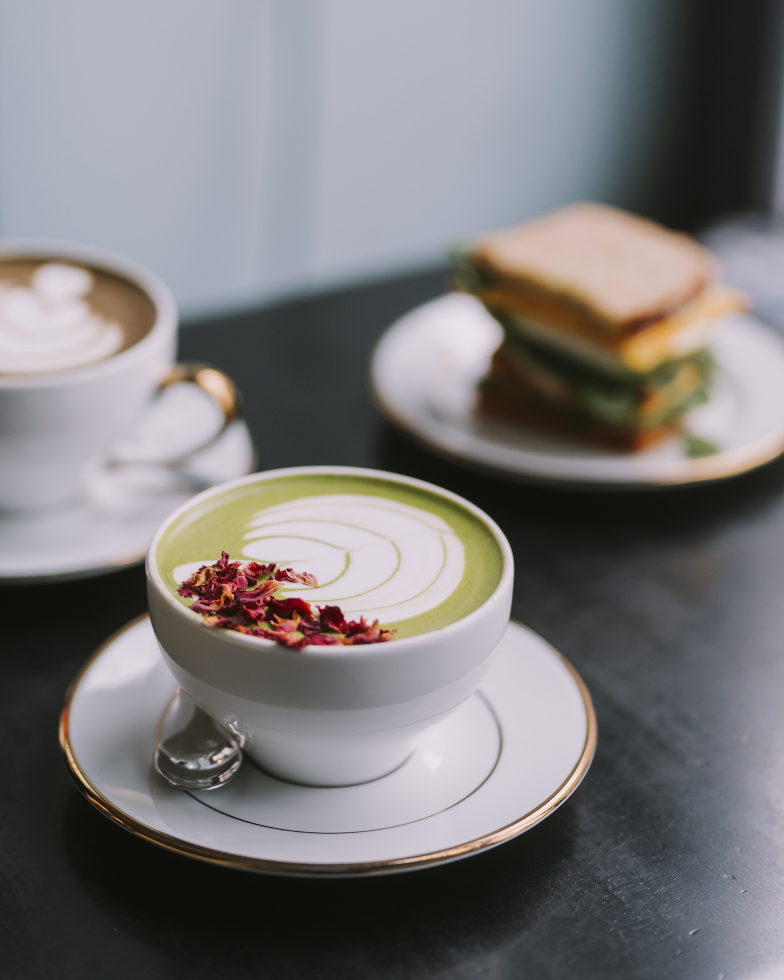 Zielona herbata matcha z dekoracją z suszonych kwiatów w kawiarni Stor, obok filiżanka kawy i kanapka.