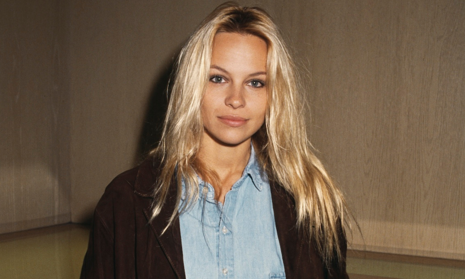 Pamela Anderson, 1993 rok, uśmiecha się, w jasnej bluzie ze stójką i brązowej kurtce. Tło jasne i neutralne.