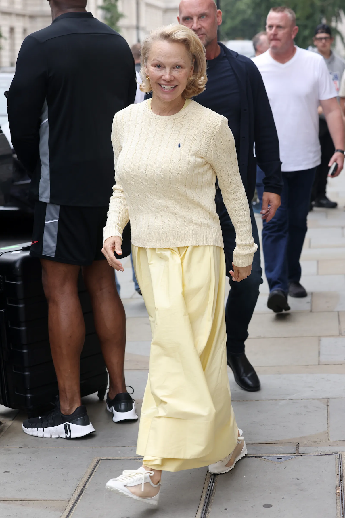 Pamela Anderson w żółtym swetrze, żółtej spódnicy i białych sneakersach Jacquemus Le Tennis, uśmiecha się na ulicy.