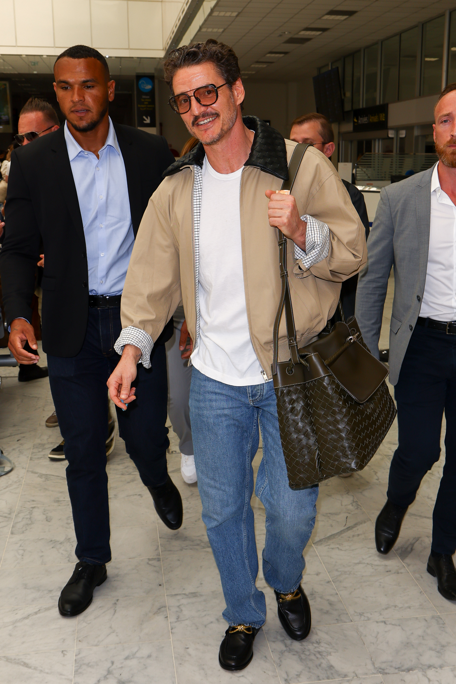 Pedro Pascal w casualowym stroju, z torbą, otoczony przez mężczyzn, na tle lotniska.