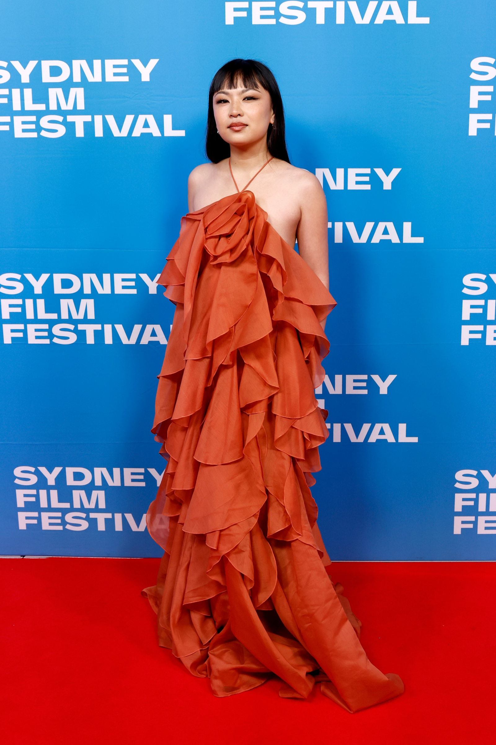 Kobieta w pomarańczowej sukience maxi w stylu boho z falbanami, pozująca na tle dla festiwalu filmowego.