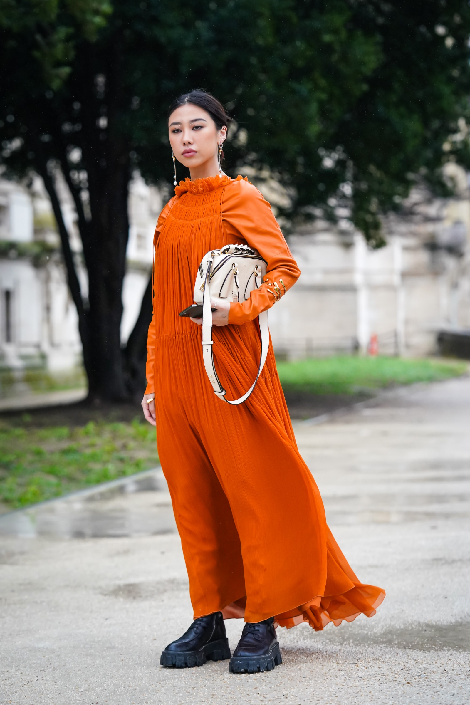 Kobieta w eleganckiej pomarańczowej sukience maxi w stylu boho, trzyma torbę, stoi na mokrej nawierzchni, w tle drzewa.
