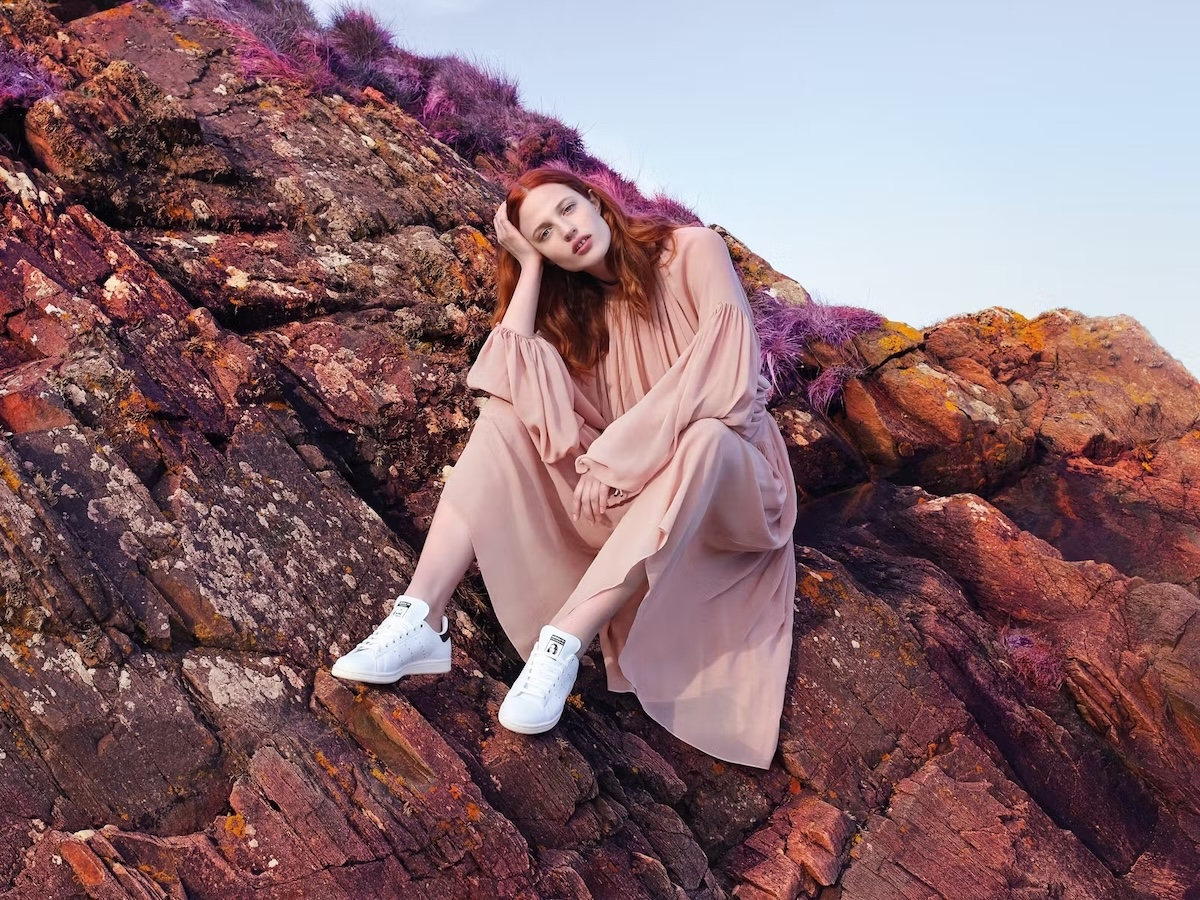 Julia Banaś w długiej, jasnej sukience siedzi na skałach, nosząc sneakersy adidas Stan Smith.