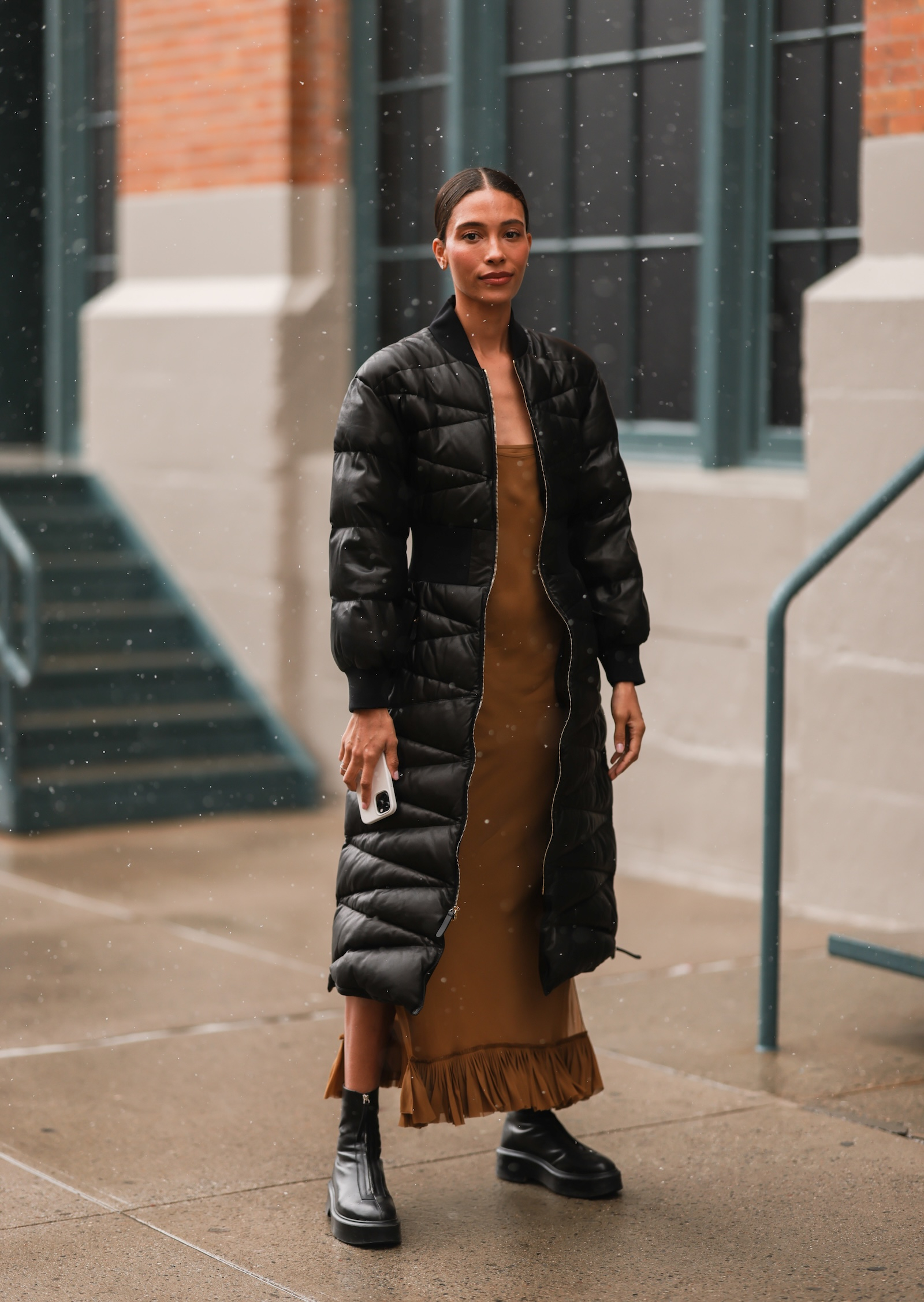 Dama w długiej pikowanej czarnej kurtce damskiej, długiej sukience i czarnych botkach stoi na ulicy.