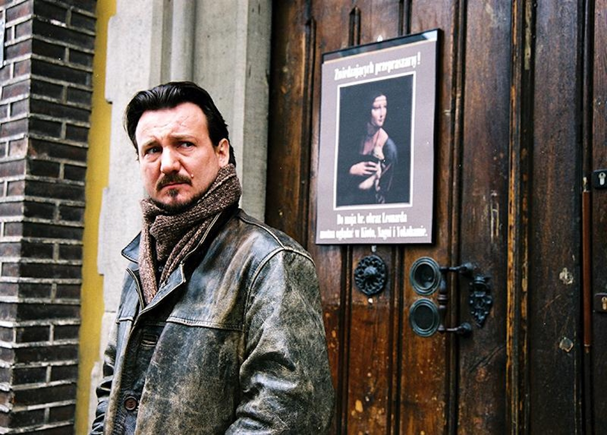 Robert Więckiewicz w filmie „Vinci” z 2004 roku stoi na ulicy, obok drzwi z plakatem.