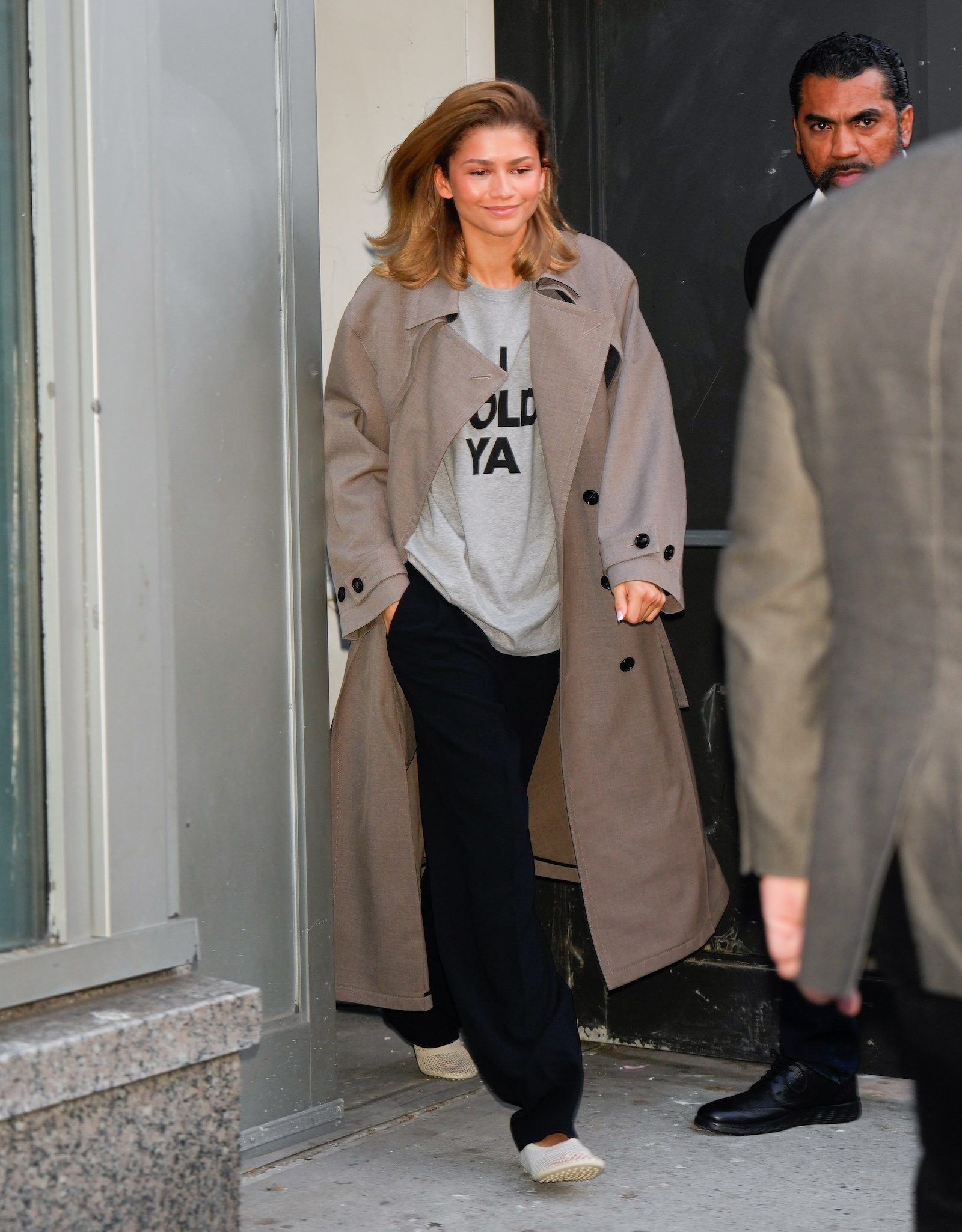 Zendaya w płaszczu, wychodząca z budynku w 2024 roku, ubrana w szary t-shirt i czarne spodnie.