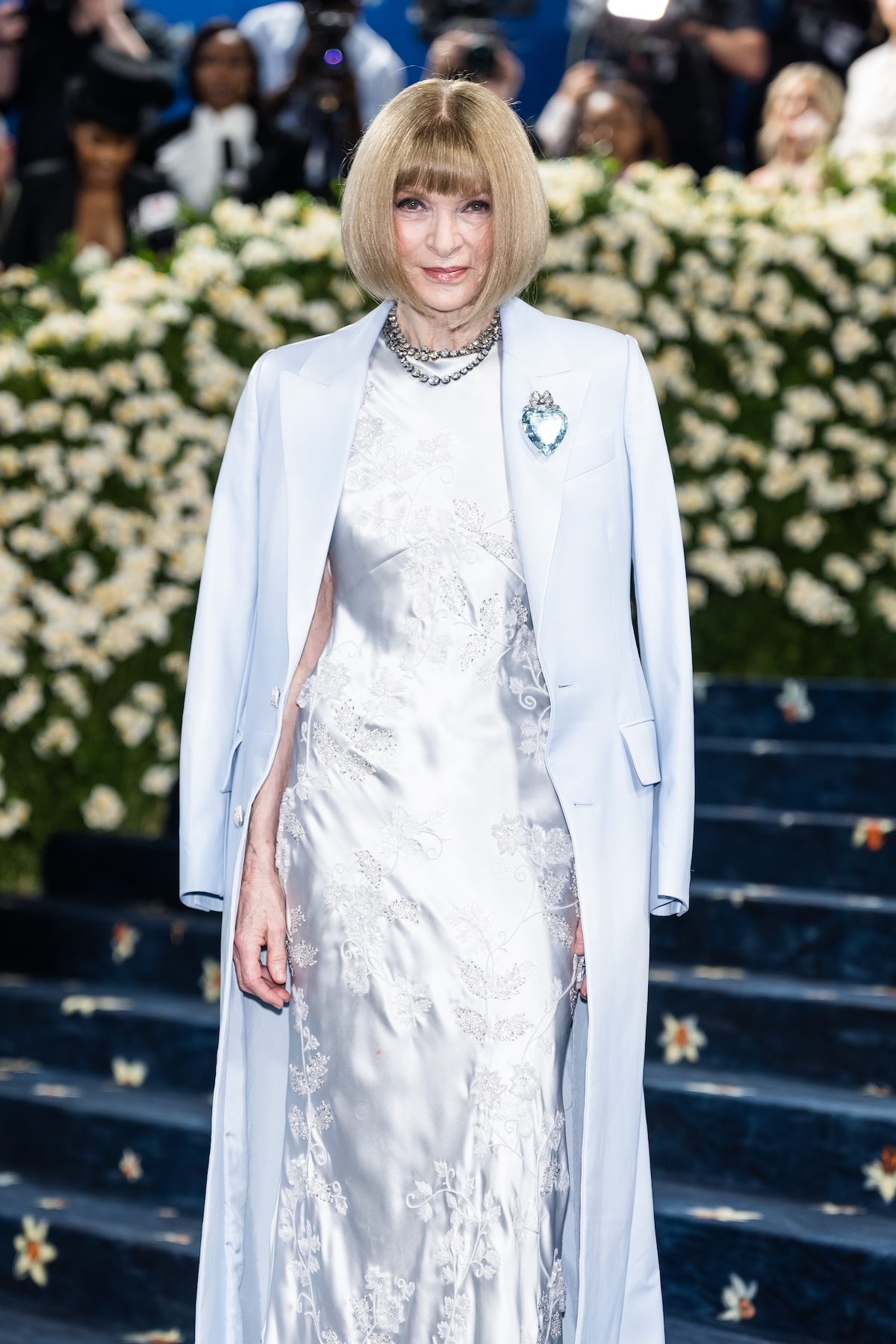 Anna Wintour na MET Gala 2025 w eleganckiej, błękitnej sukni i pelerynie, w otoczeniu kwiatów.