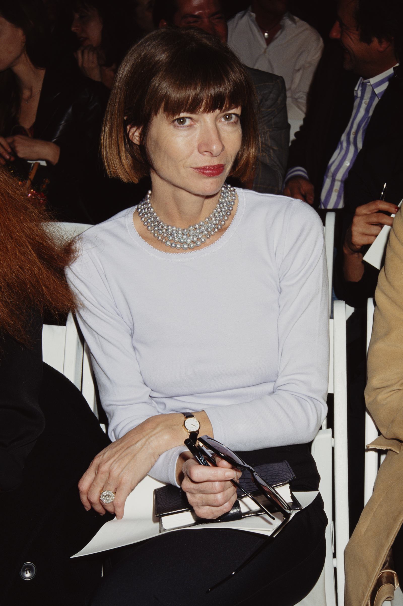 Anna Wintour, 1997 rok, siedzi w publiczności, ubrana w jasną bluzkę, z perłowym naszyjnikiem i okularami w ręku.