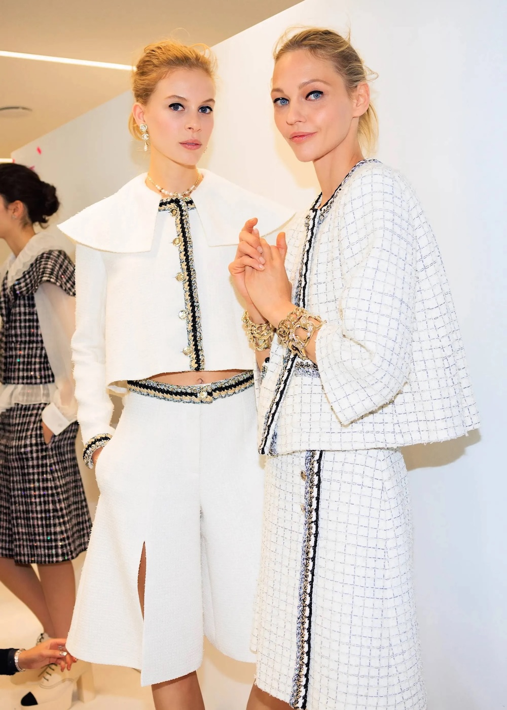 Dwie modelki w białych bermudach i eleganckich bluzkach, prezentujące stylowe zestawienia w jasnym otoczeniu.