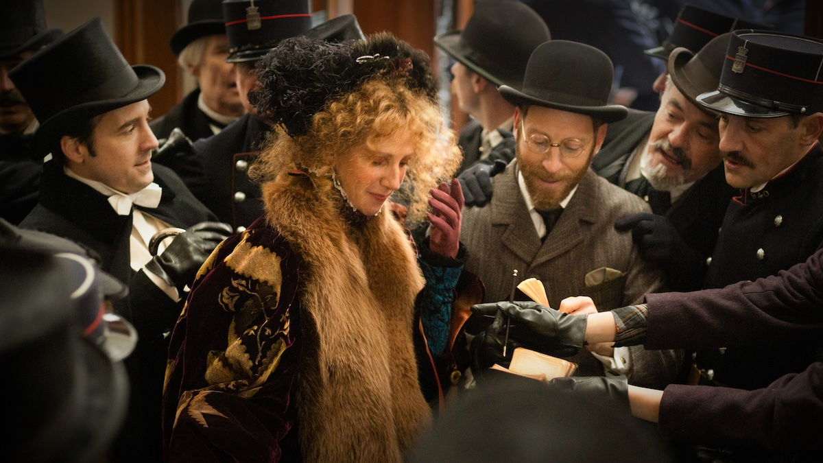 Sandrine Kiberlain jako Sarah Bernhardt w filmie „Boska Sarah Bernhardt”, otoczona mężczyznami w eleganckich strojach.