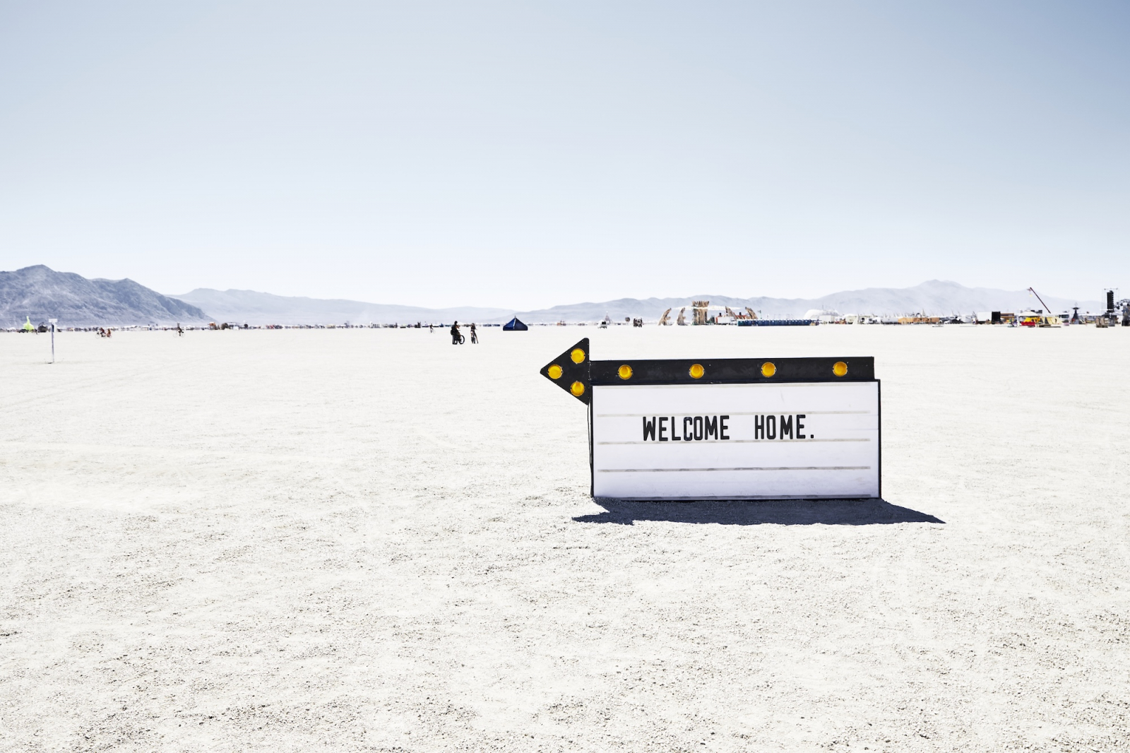 Tablica z napisem "WELCOME HOME" na pustyni podczas festiwalu Burning Man, w tle widoczne namioty i góry.