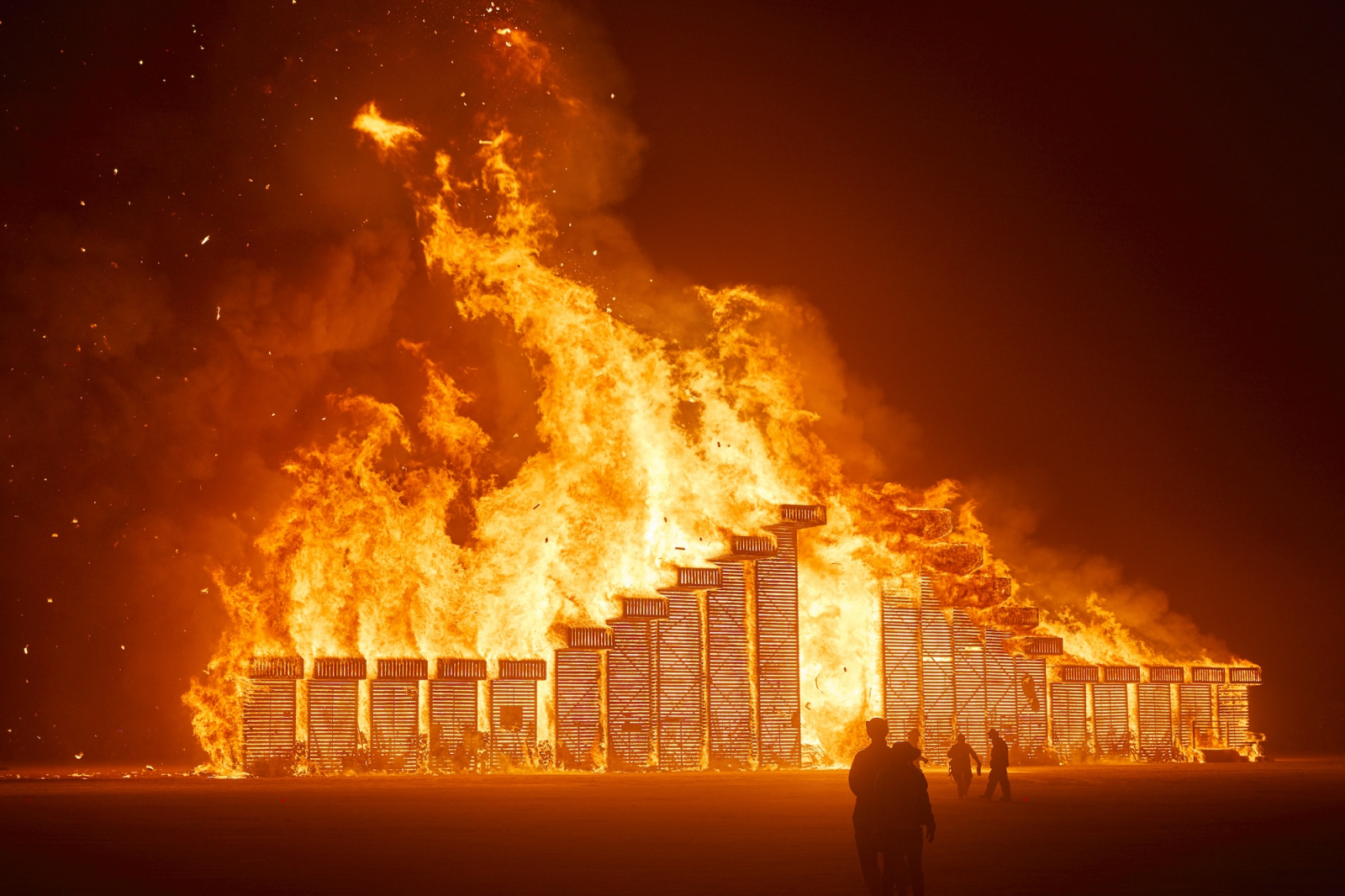 Festiwal Burning Man, ogień przetacza się przez ogromną strukturę, otaczając postacie w ciemności.