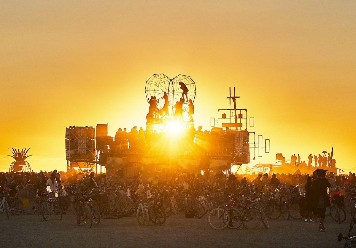 Zachód słońca na festiwalu Burning Man, z postaciami na podwyższeniu i tłumem ludzi wokół rowerów.