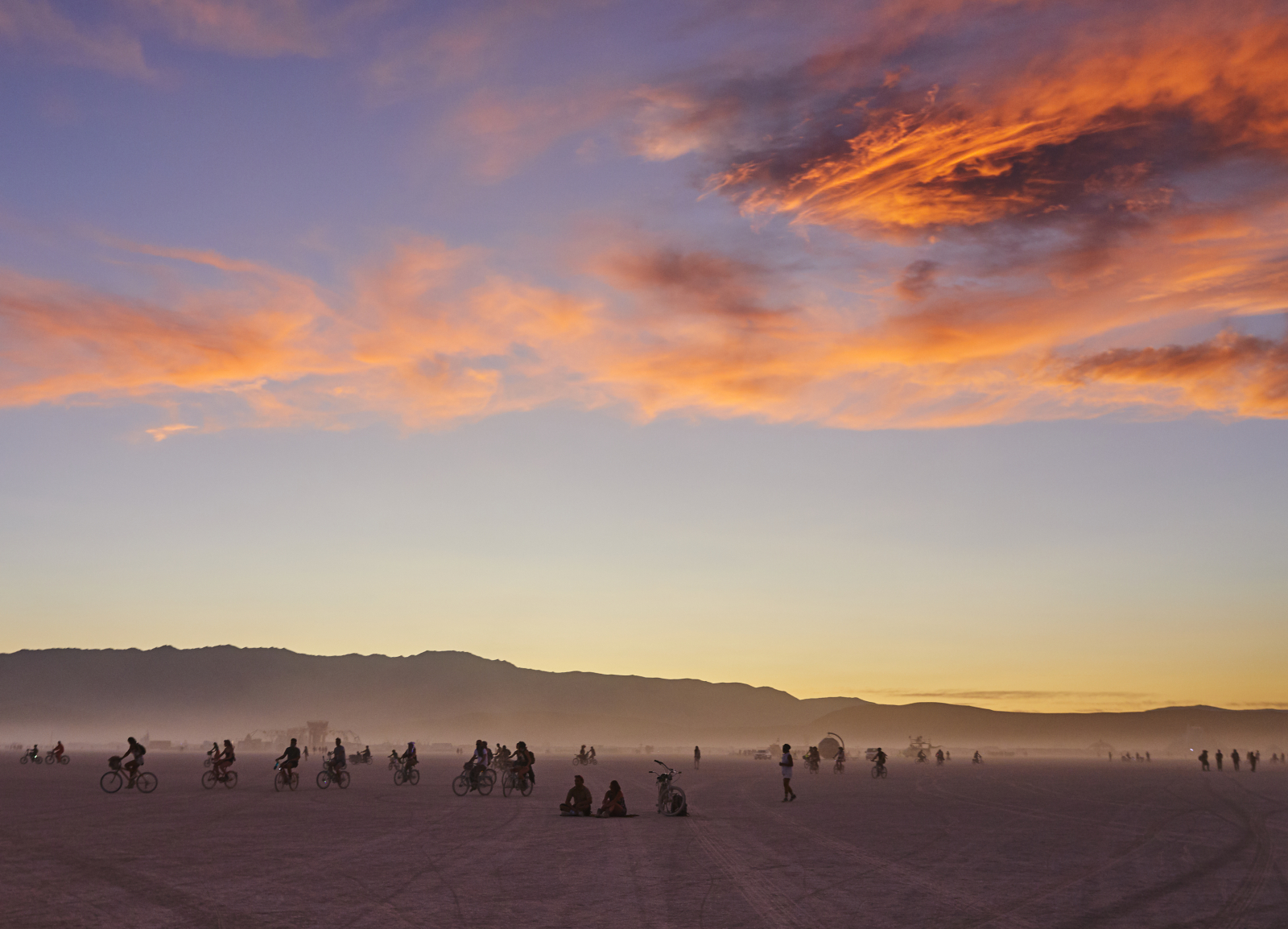 Festiwal Burning Man z ludźmi na rowerach w pustynnym krajobrazie przy zachodzie słońca.