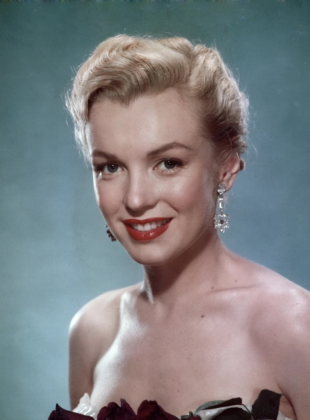 Marilyn Monroe z lat 50-tych, z eleganckim makijażem, srebrnymi kolczykami i kwiatami w dekolcie.