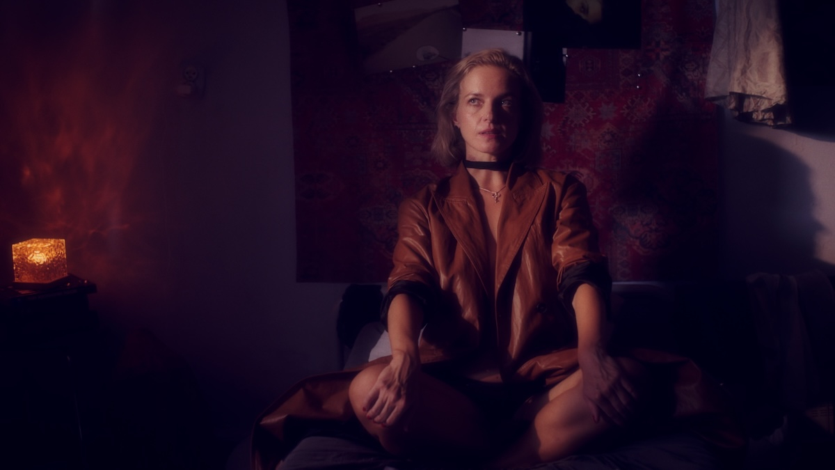 Marta Nieradkiewicz w filmie „Trzy miłości”, siedzi na łóżku w stylowej brązowej pelerynie, w nastrojowym świetle.