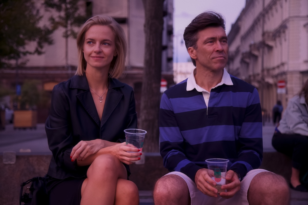 Marta Nieradkiewicz i Marcin Czarnik w filmie „Trzy miłości”, siedzą na ławce w miejskim otoczeniu, trzymając kubki.