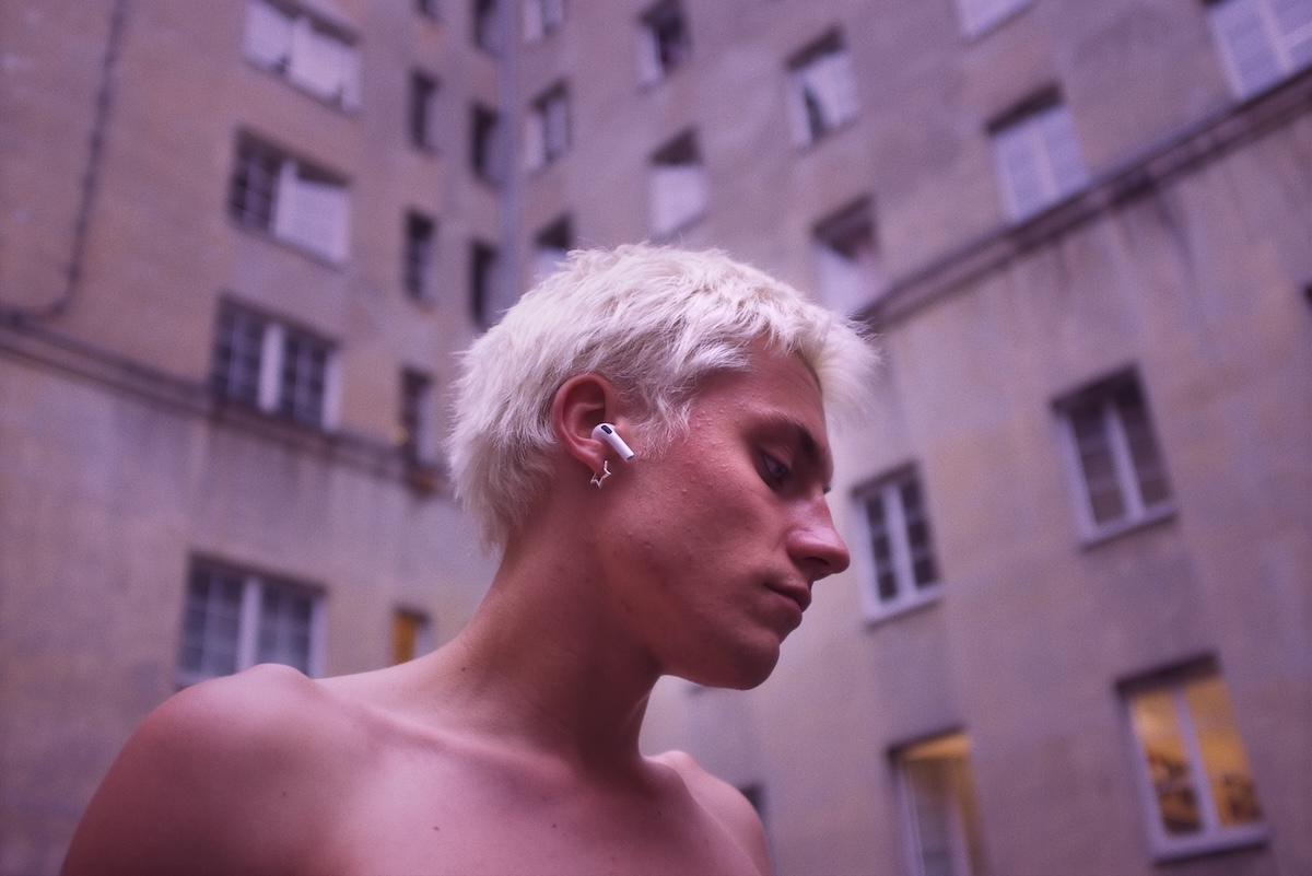 Mieszko Chomka w filmie „Trzy miłości”, spoglądający w dół, z krótkimi, blond włosami i słuchawką w uchu.